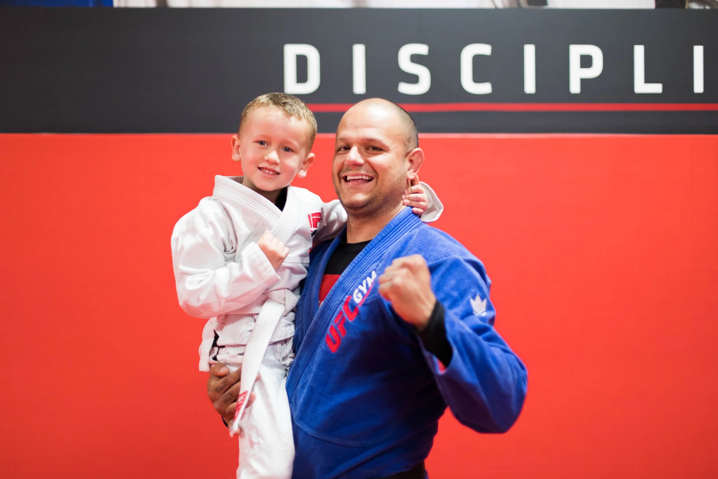 A young boy wearing a white Brazilian Jiu-Jitsu gi with a black belt, smiling and posing with an adult man in a blue Brazilian Jiu-Jitsu gi with a black belt, in front of a sign that reads 'Discipline'. Both appear happy and confident.