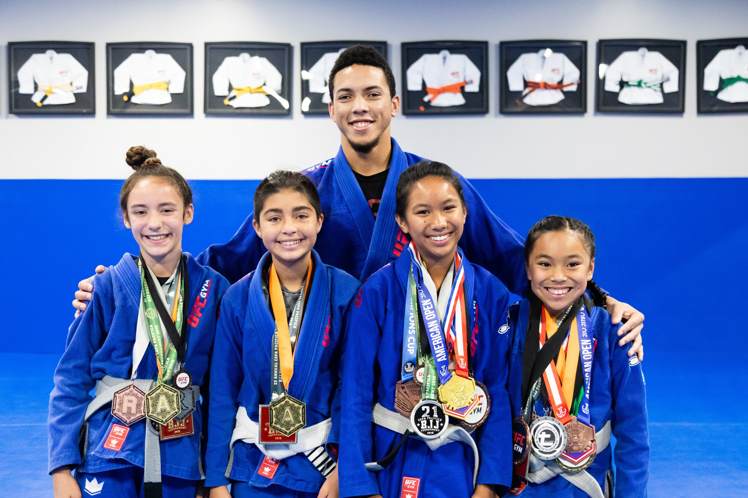 Group of young girls and their adult instructor in Brazilian Jiu-Jitsu uniforms, smiling and showing medals after a competition, with framed gi uniforms on the wall in the background.