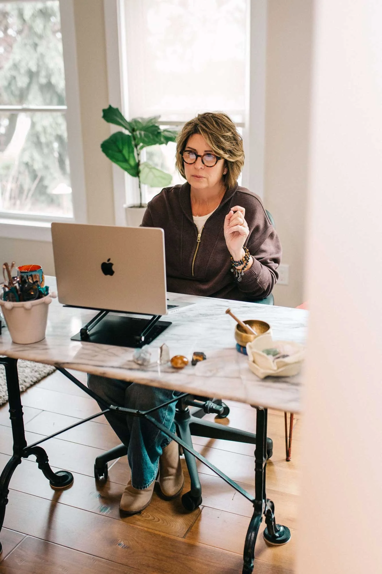 A woman with glasses, short wavy hair, wearing a brown jacket, sits at a white table in a well-lit room, working on a laptop with an Apple logo. She has bracelets on her wrist and tan boots. The table has various small items, including stones, a container with pens, and a bowl. Behind her is a window with blinds and a potted plant.