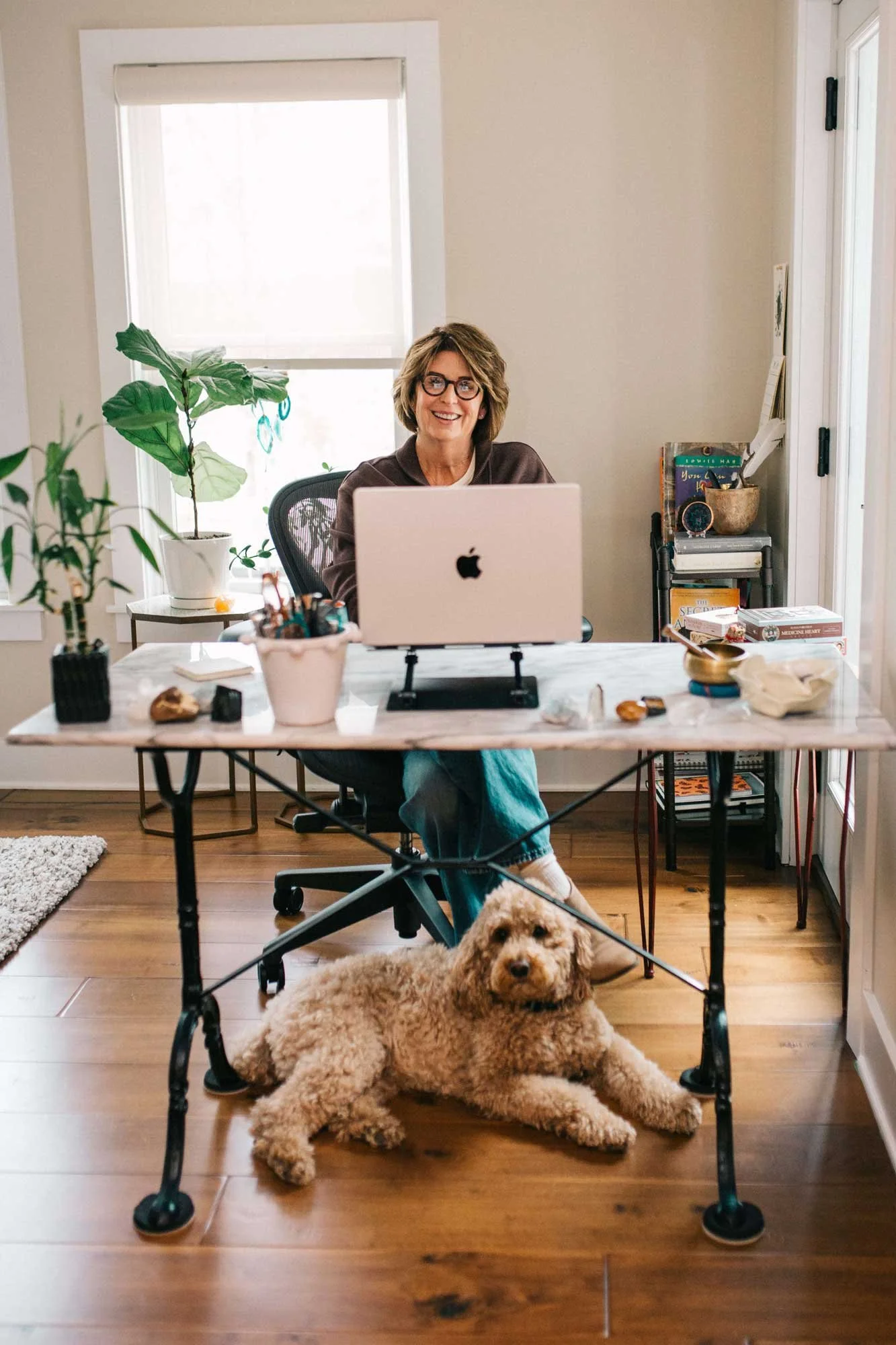 A woman with glasses sitting at a desk with a laptop, smiling. A dog is lying on the wooden floor underneath the desk. The room has a window with a shade, a bookshelf with books and decorative items, and various potted plants.