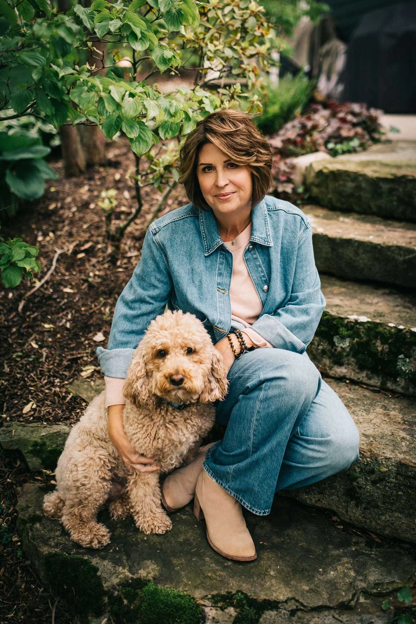 A woman with short brown hair wearing a denim jacket and beige boots sitting on stone steps in a garden, holding a fluffy beige poodle dog.
