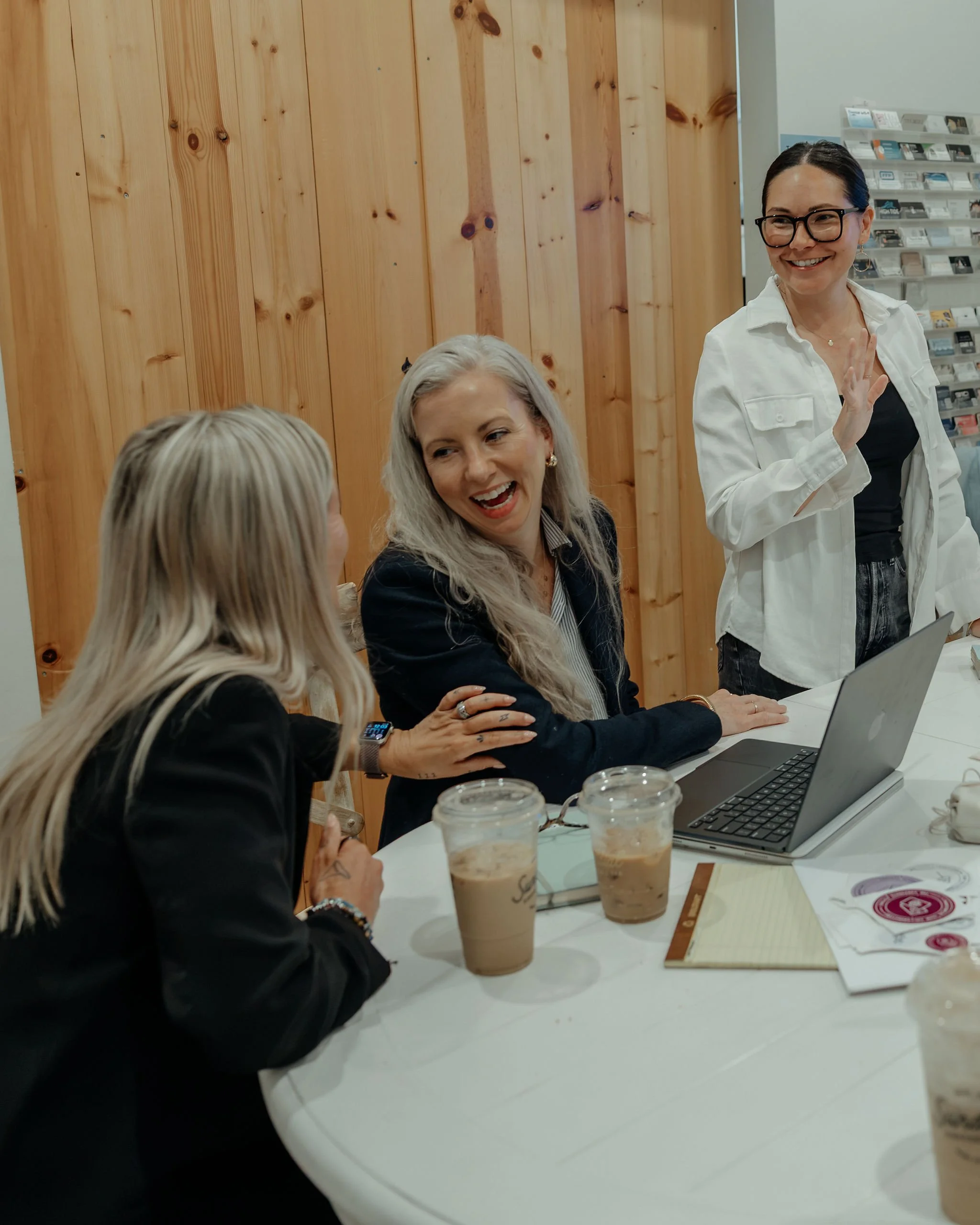 Women sitting at a white table in a coffee shop, smiling and talking, with three iced coffee drinks in front of them, and a woman standing with a white shirt and glasses waving.