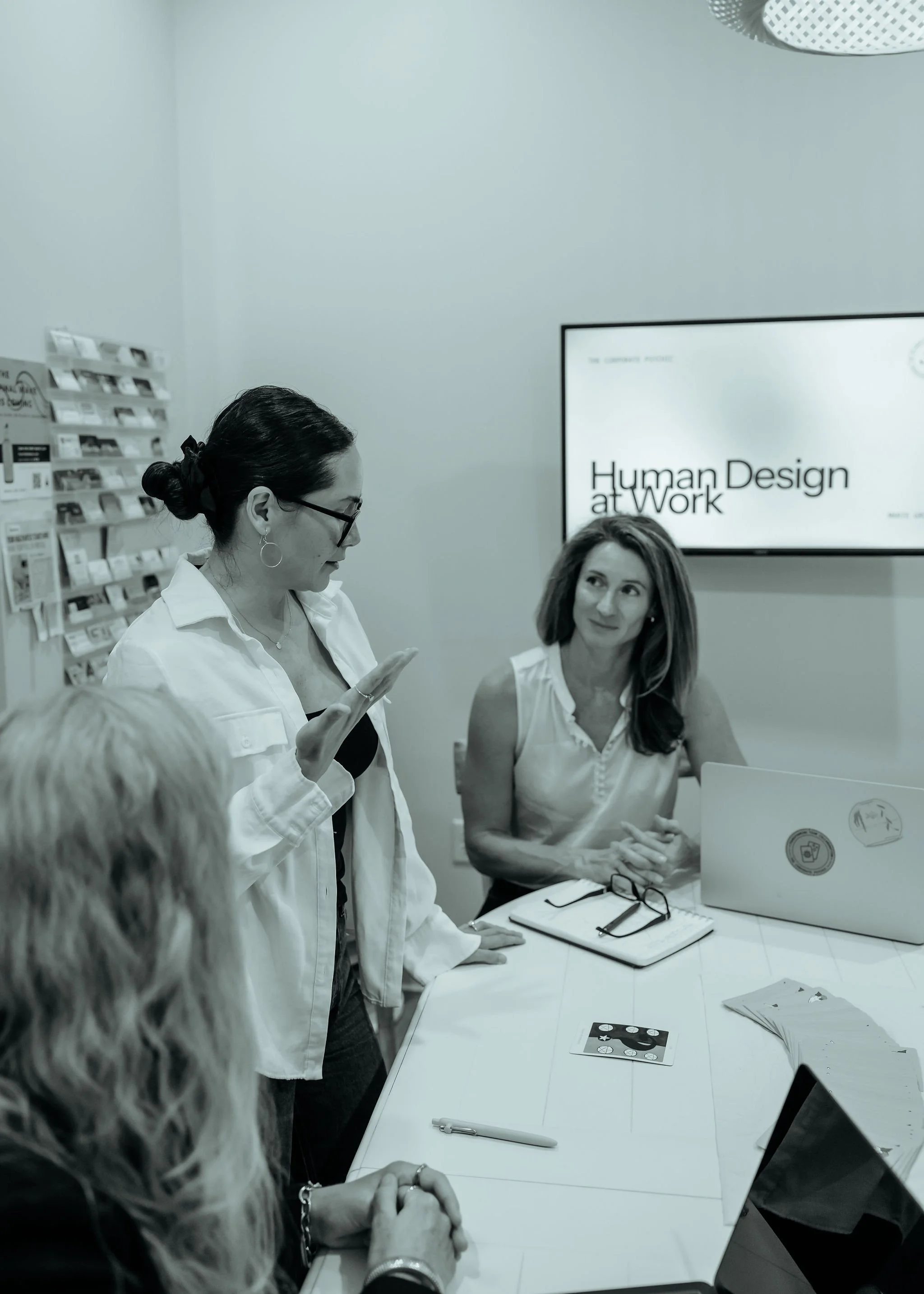 Group of women in a meeting room, one woman standing and speaking, others seated, with a presentation slide titled 'Human Design at Work' on the wall.
