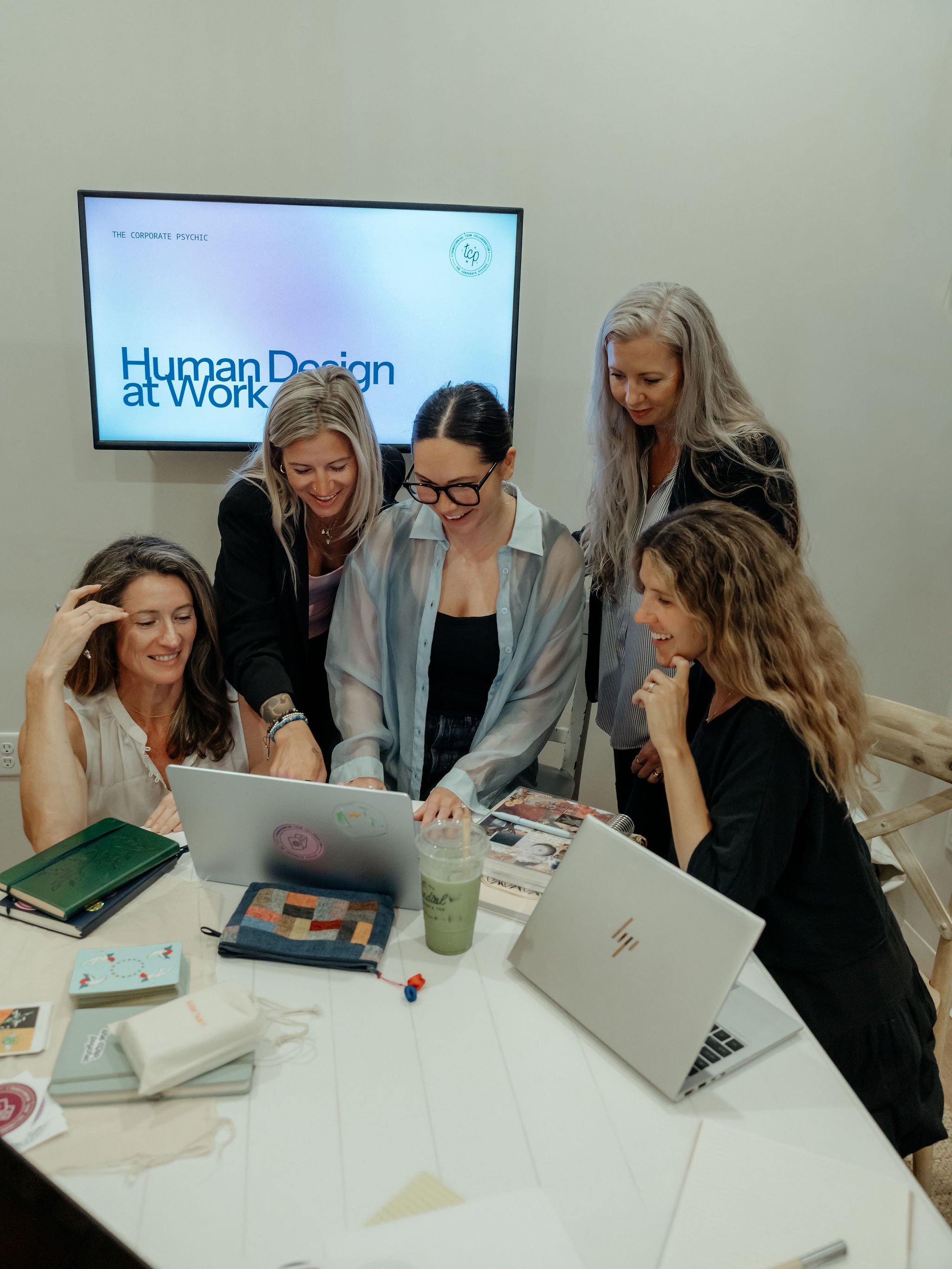 A group of women gathered around a table with laptops, magazines, notebooks, and drinks, looking at a laptop and engaging with each other in an indoor setting.