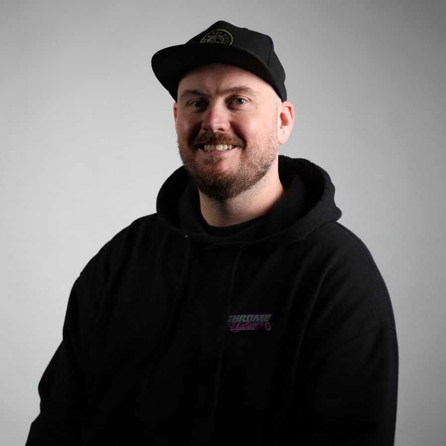 A smiling man with a beard and mustache wearing a black hoodie and a black cap, standing against a plain light gray background.