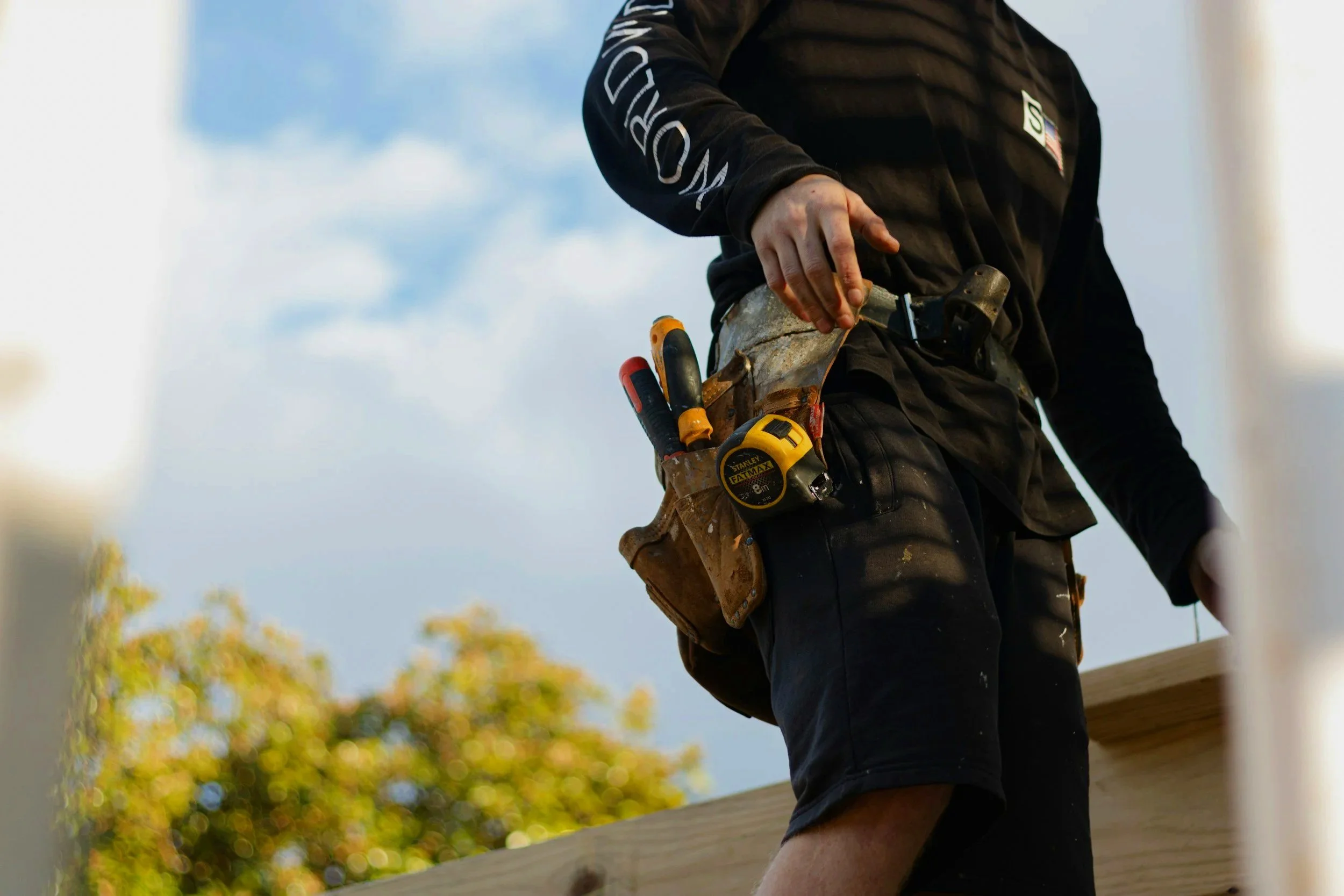 A person wearing black work clothes with a belt and tools, including a tape measure and a screwdriver, in a leather holster, working on a construction or woodworking project outdoors during daytime.