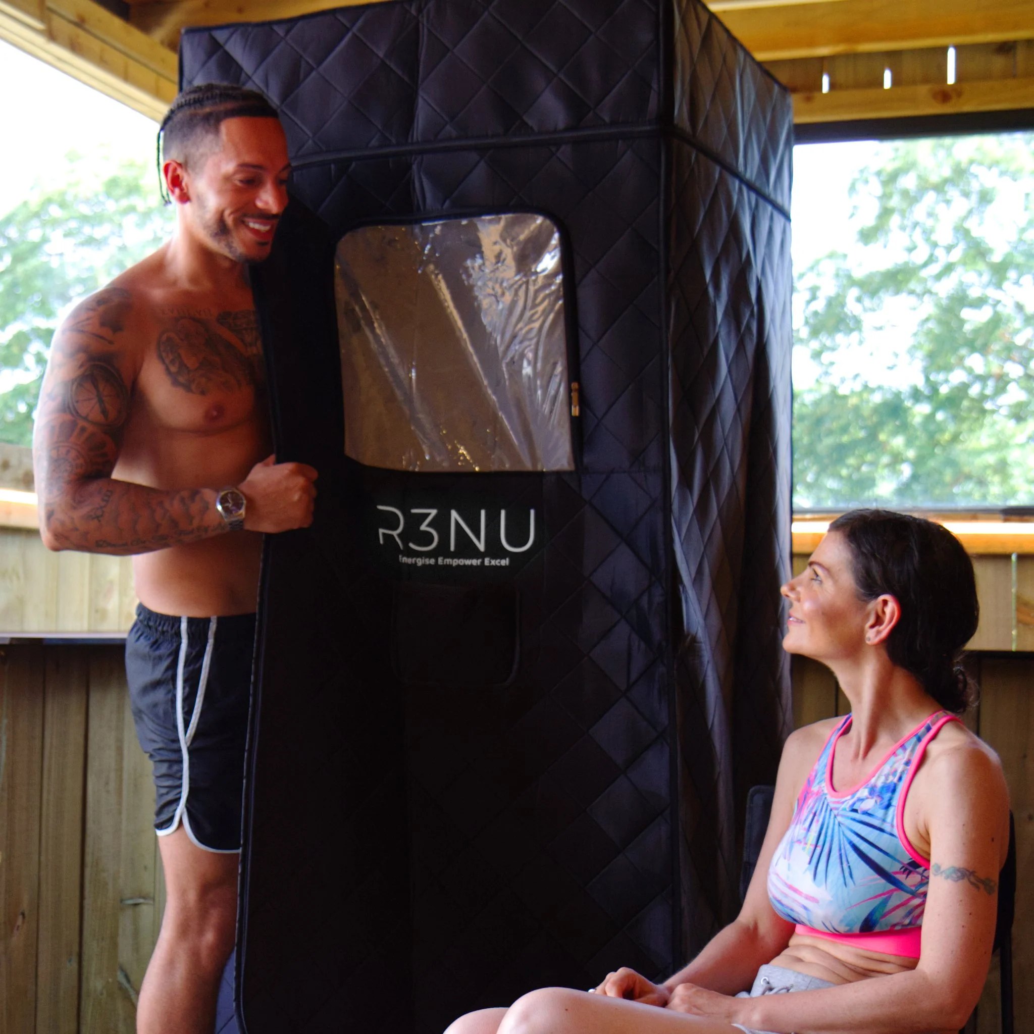 A shirtless man with tattoos and a woman with dark hair sitting outdoors; large black quilted object, possibly a speaker or equipment, is visible in the background.