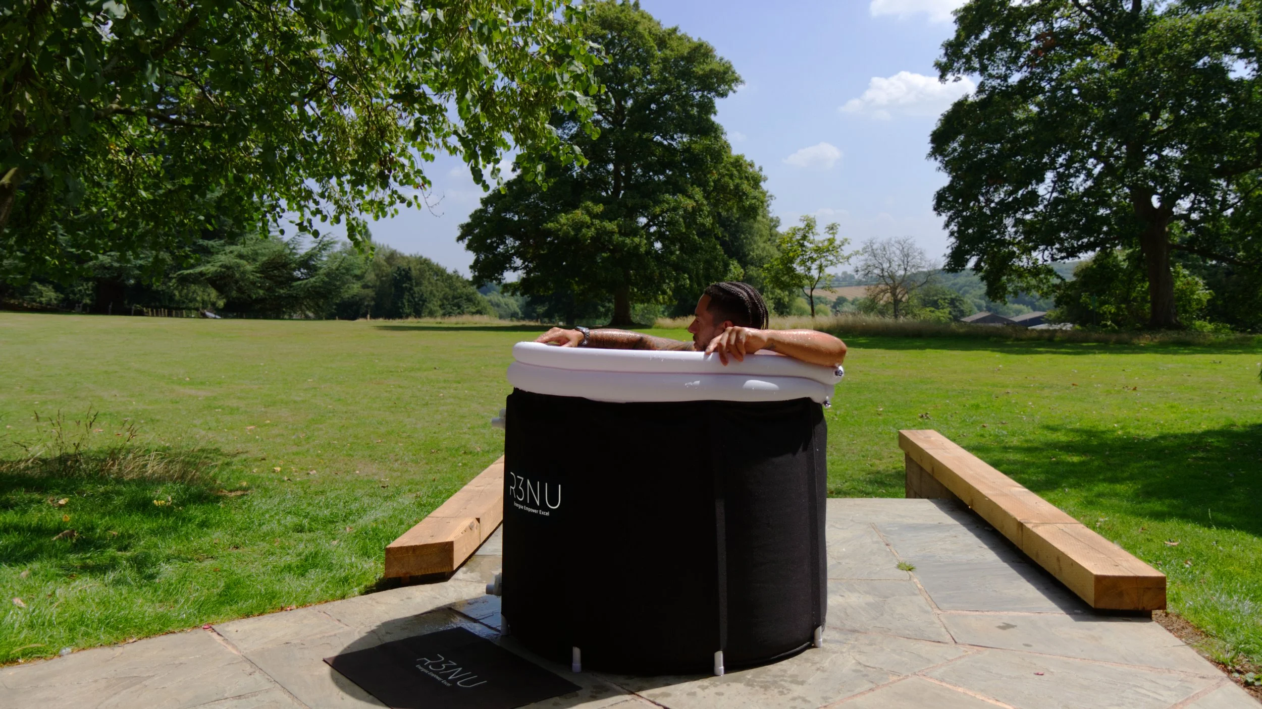 A person with dark hair relaxing in an inflatable hot tub outdoors, surrounded by greenery and trees on a sunny day.