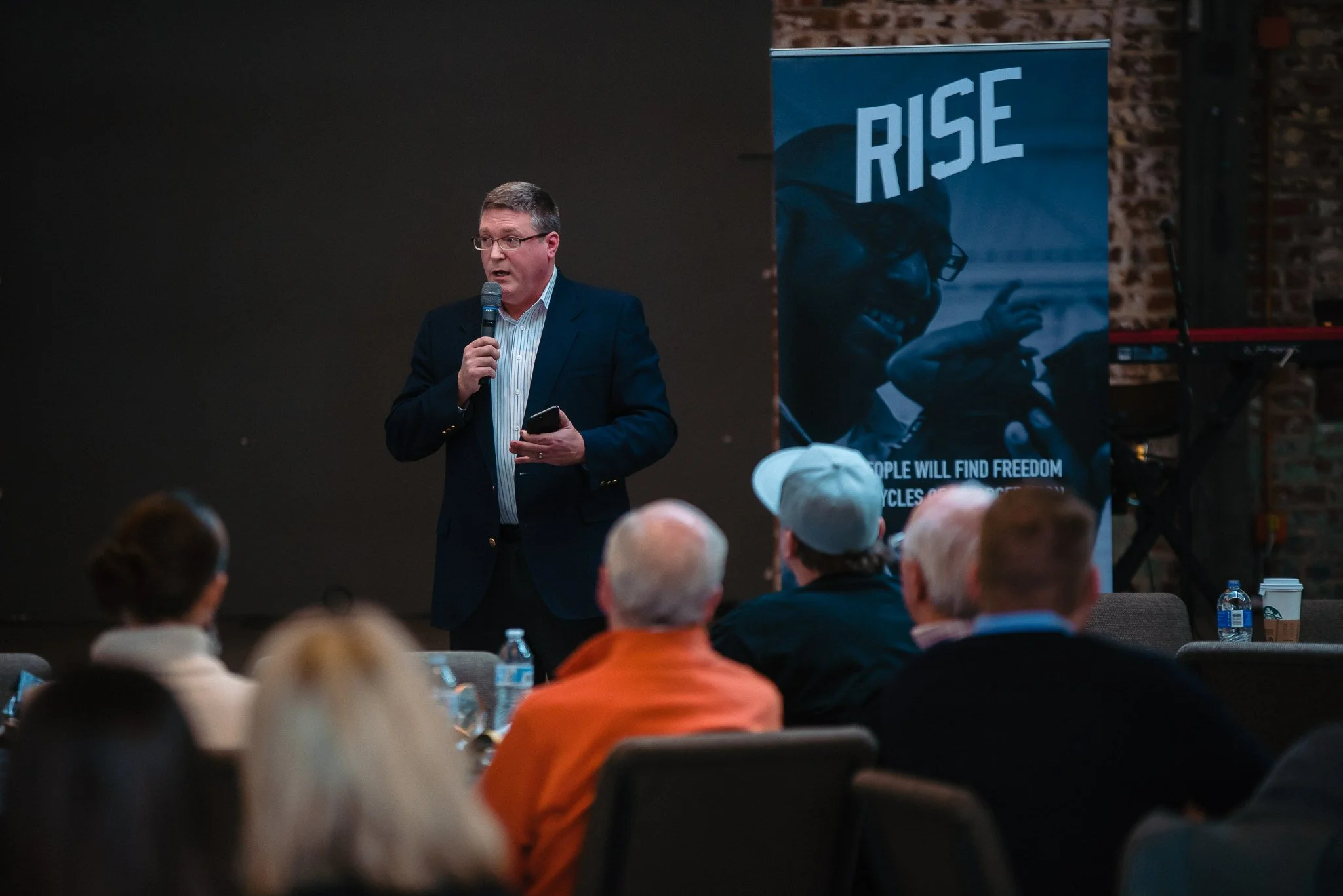A man in a dark suit and striped shirt giving a presentation with a microphone in a room with an audience. A large blue banner with the word 'RISE' and a smiling person with glasses is visible in the background.
