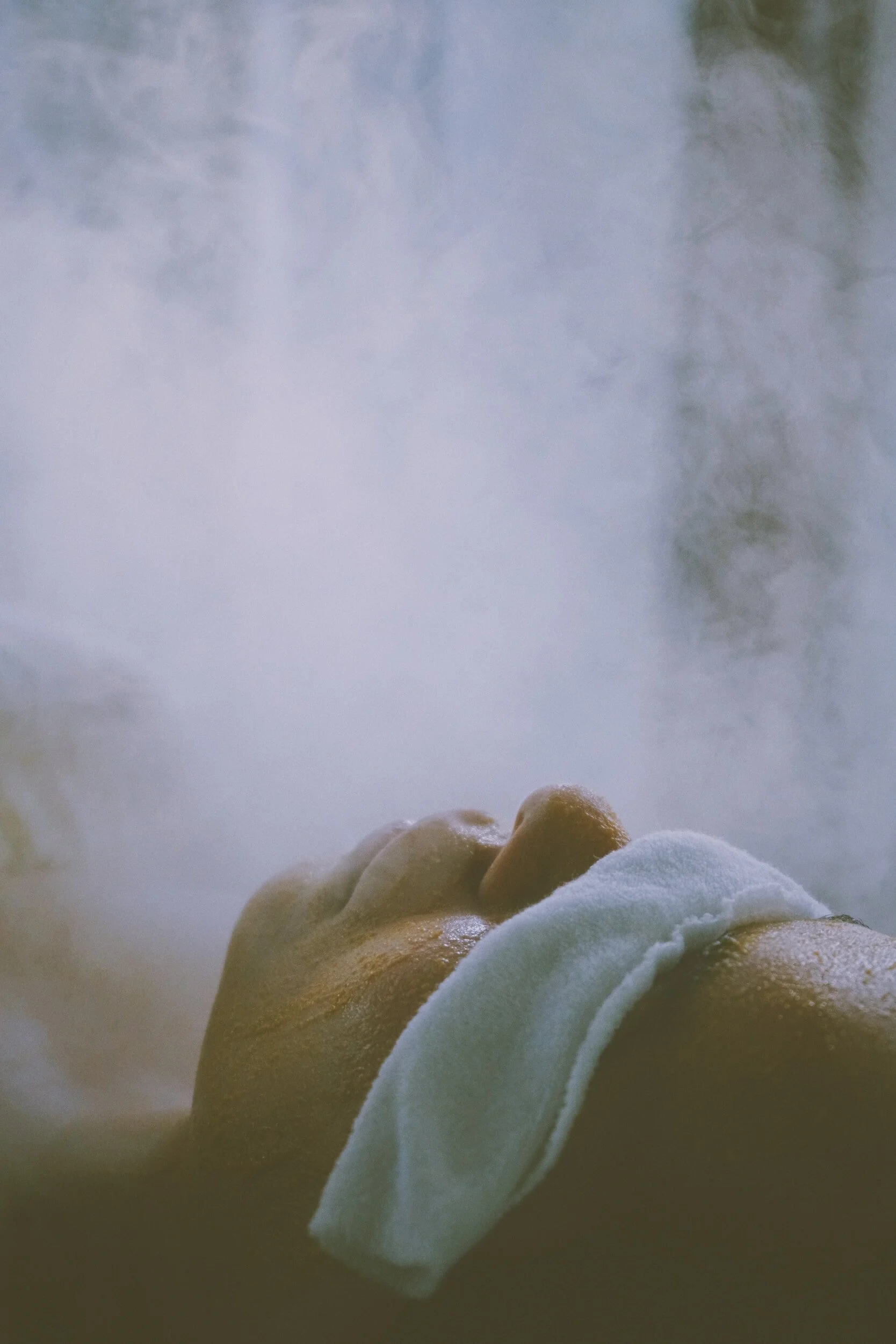 Person lying down with face covered by a towel, surrounded by steam, indicating a spa or sauna setting.
