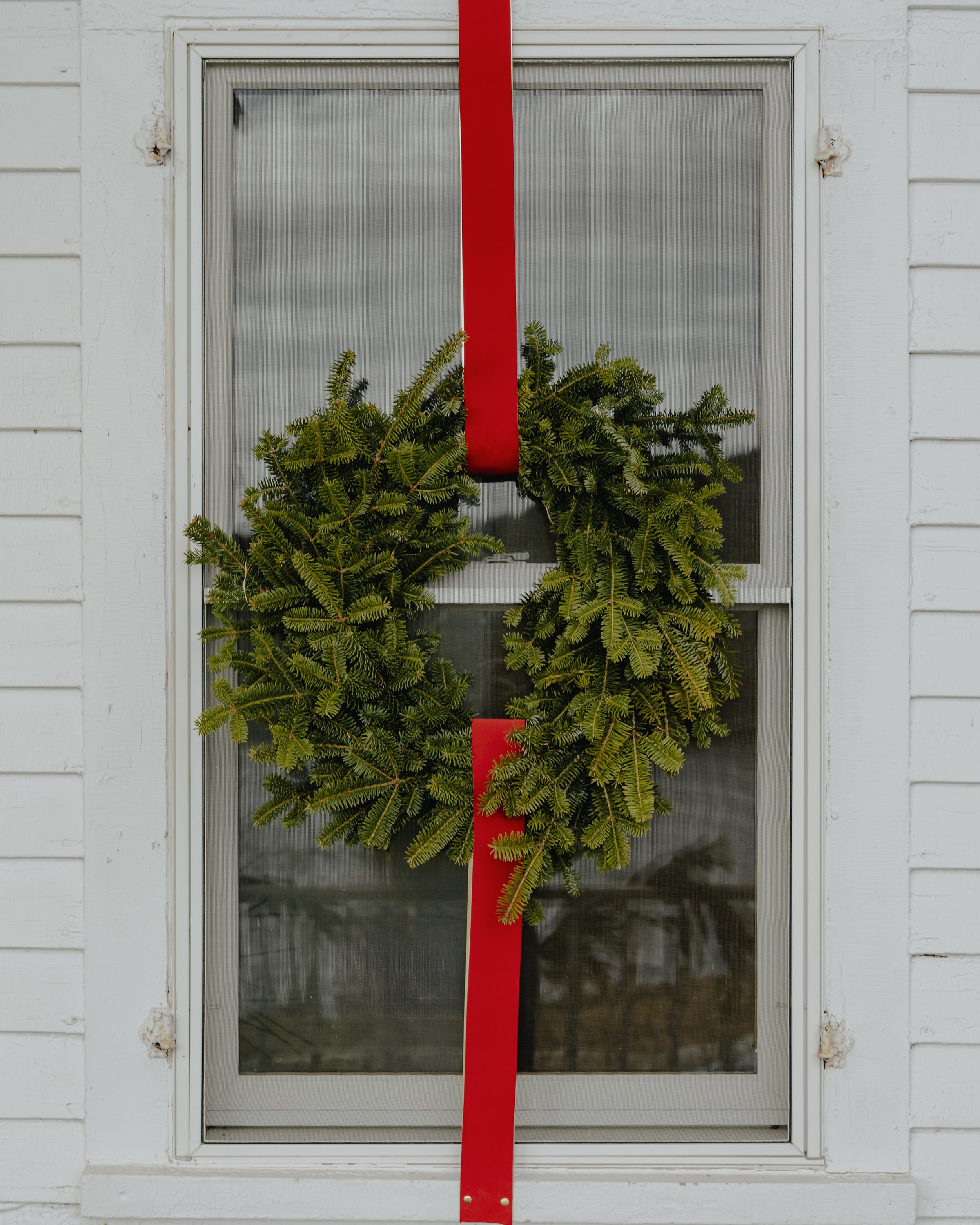 Green holiday wreath with a red ribbon hanging on a window screen in front of a white wooden house.