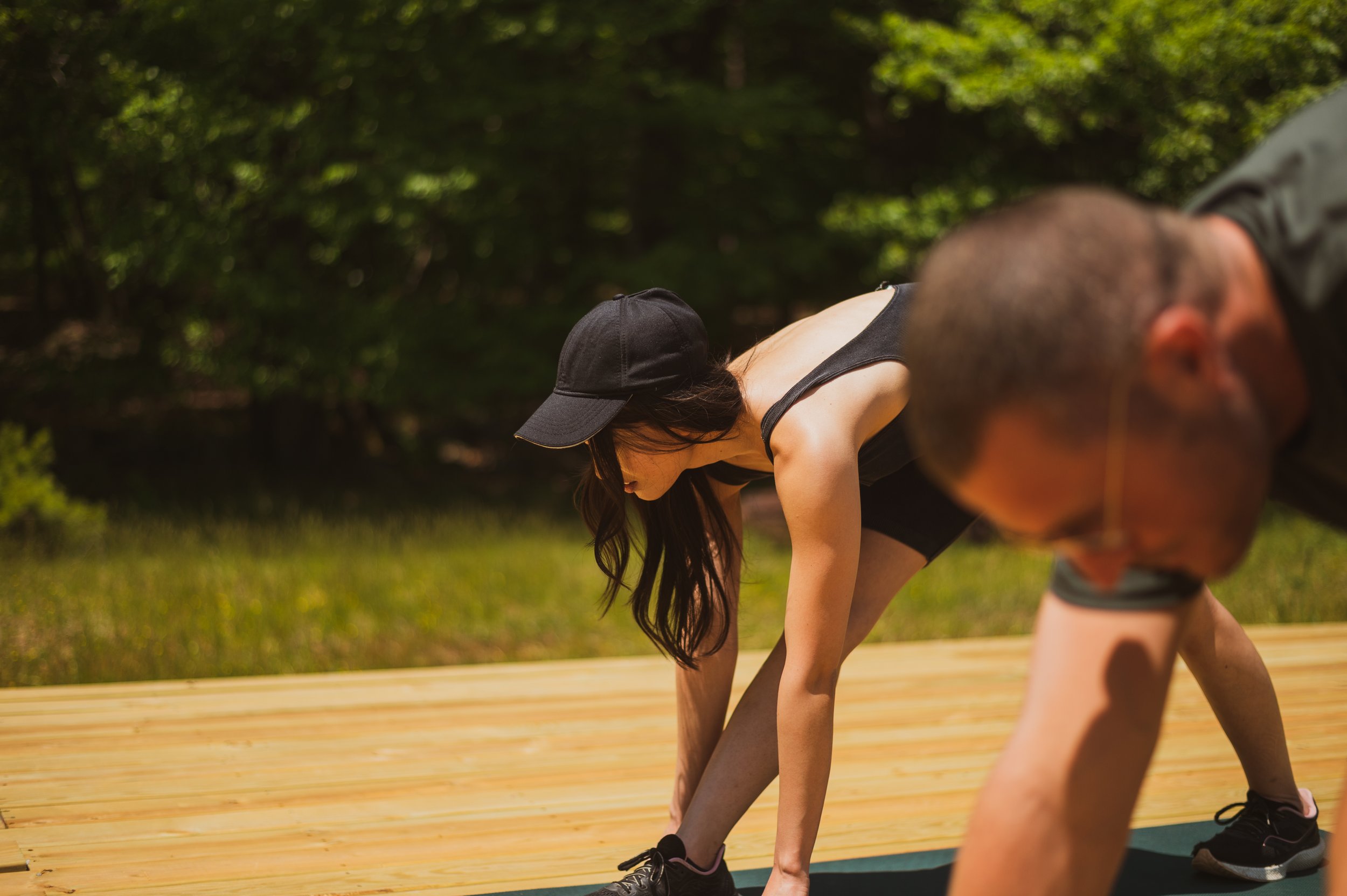 Two people stretching outdoors on a wooden deck, with a woman in a black cap and tank top bending down, and a man in a gray T-shirt also bending forward.