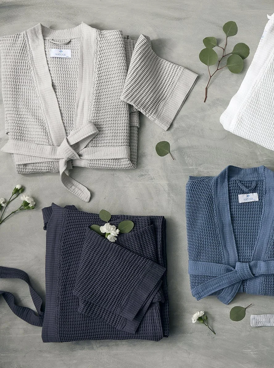 Three folded waffle-knit robes in gray, navy, and blue arranged on a light surface with green eucalyptus leaves and white flowers scattered around.