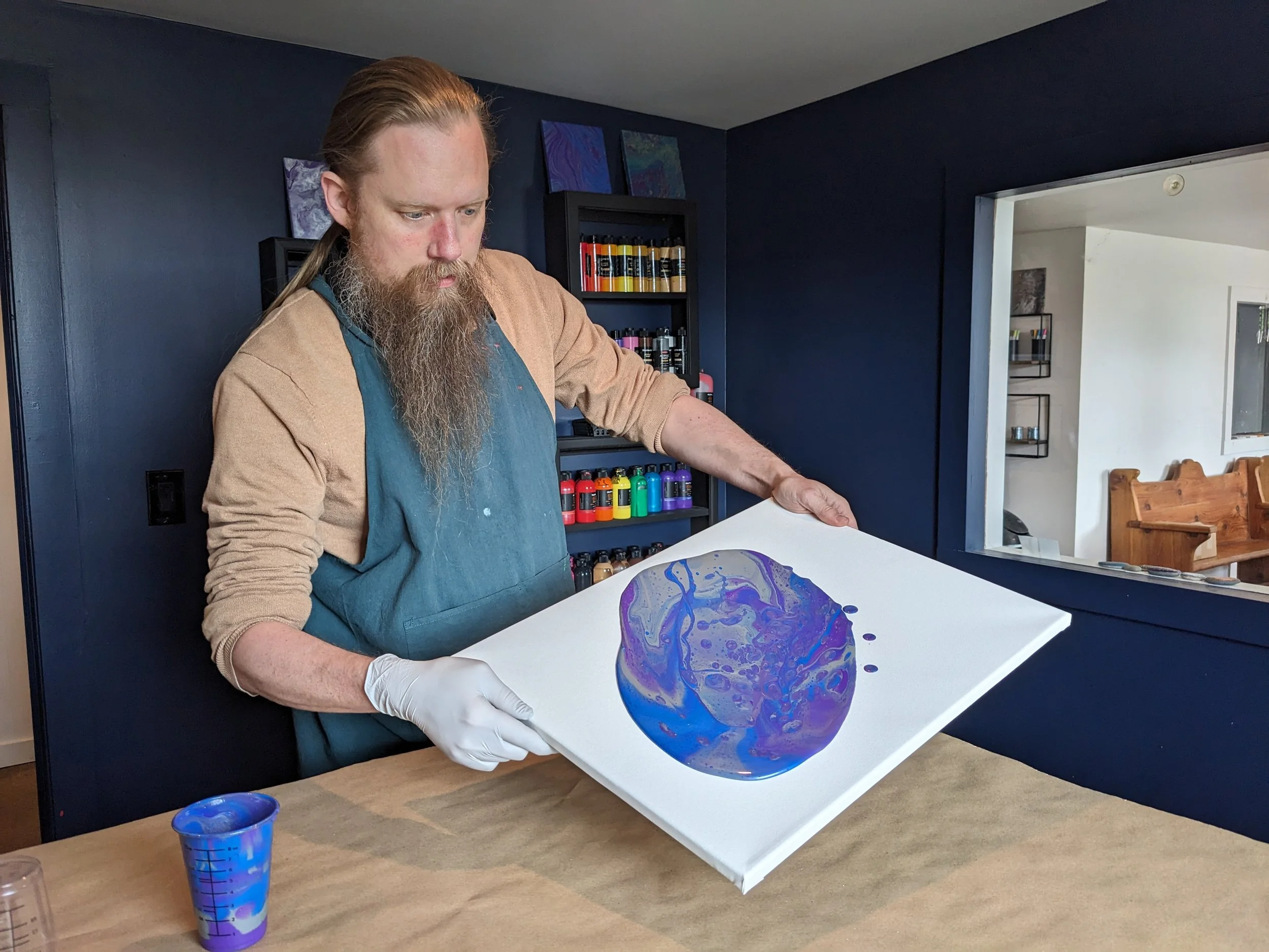 A man with long red hair and beard wearing a blue apron and beige sleeves is holding a canvas with abstract blue and purple swirling paint in an art studio.