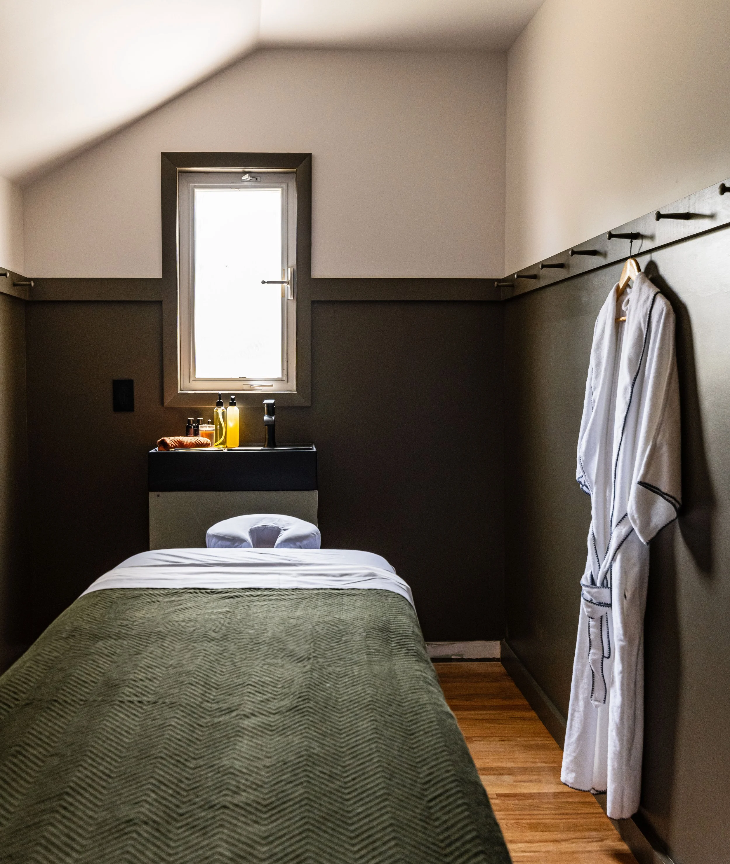 A small massage room with a single massage table covered with a green blanket, a small window on the wall, and a bathrobe hanging on hooks on the right wall.
