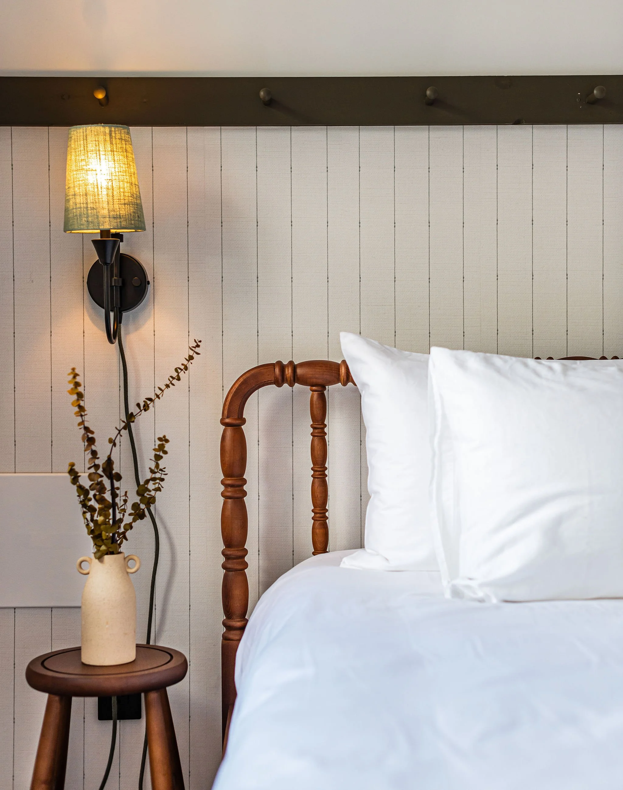 Close-up of a cozy bedroom with a wooden bed frame, white bedding, a bedside table with a beige vase holding dried branches, a wall-mounted lamp, and a wood-paneled wall.