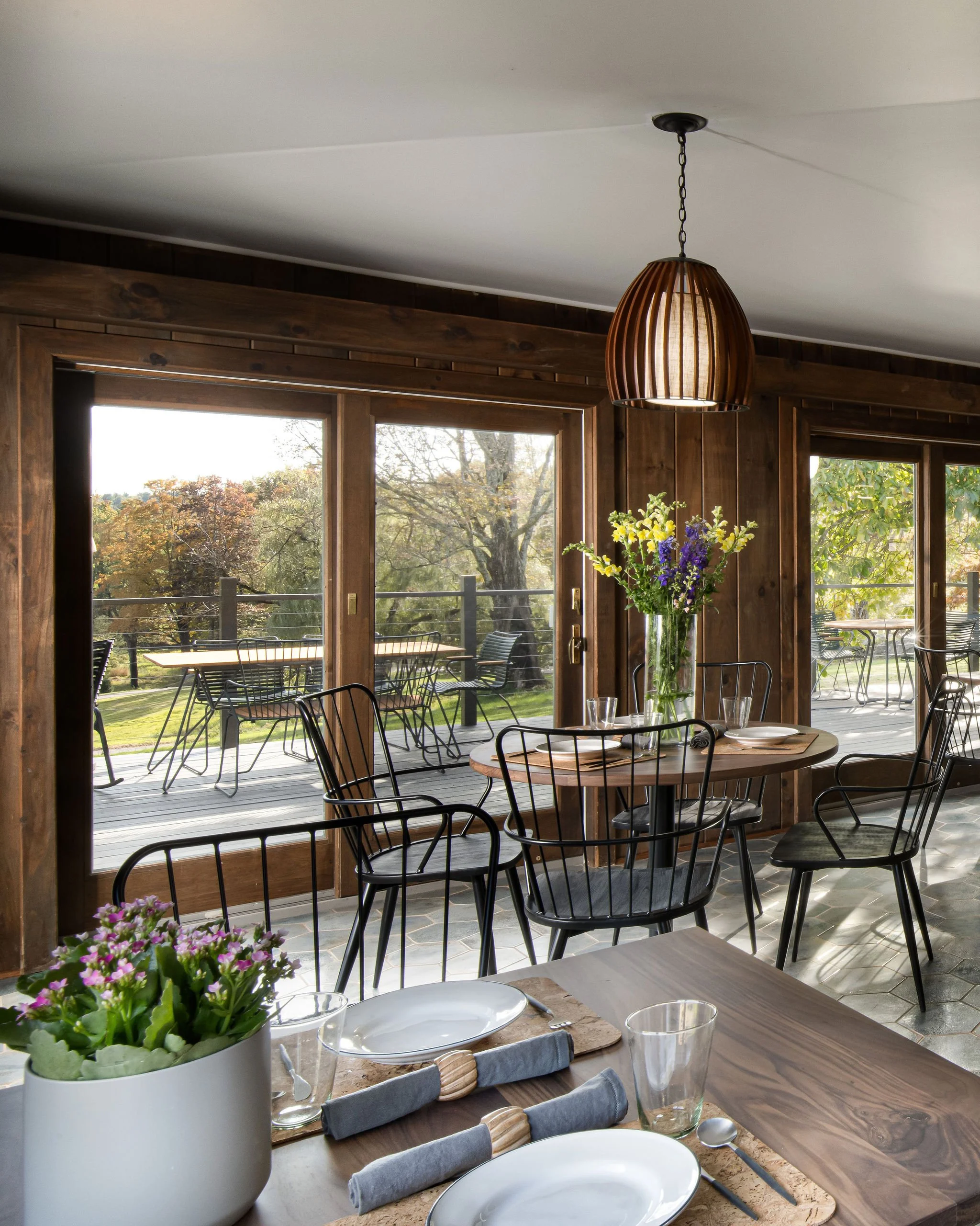 A dining area inside with wooden walls, a round wooden table set with plates, glasses, and napkins, and a large glass vase with purple and yellow flowers as a centerpiece. Large sliding glass doors open to a deck with outdoor seating and view of trees.