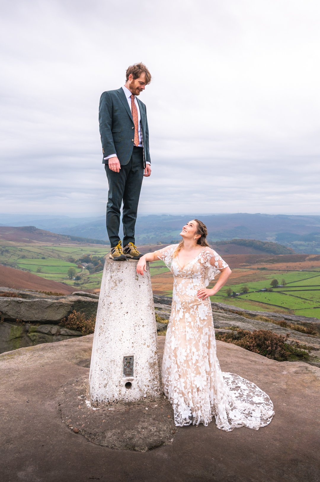 A man in a suit standing on top of a stone marker, looking down at a woman in a wedding dress standing on the ground, with a scenic countryside background, peak district wedding photography. 
