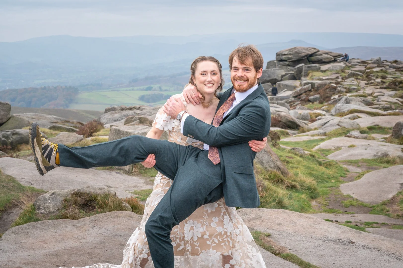 A smiling man in a suit holding a woman in a floral dress in a mountain landscape with rocks and distant hills, peak district wedding photography. 