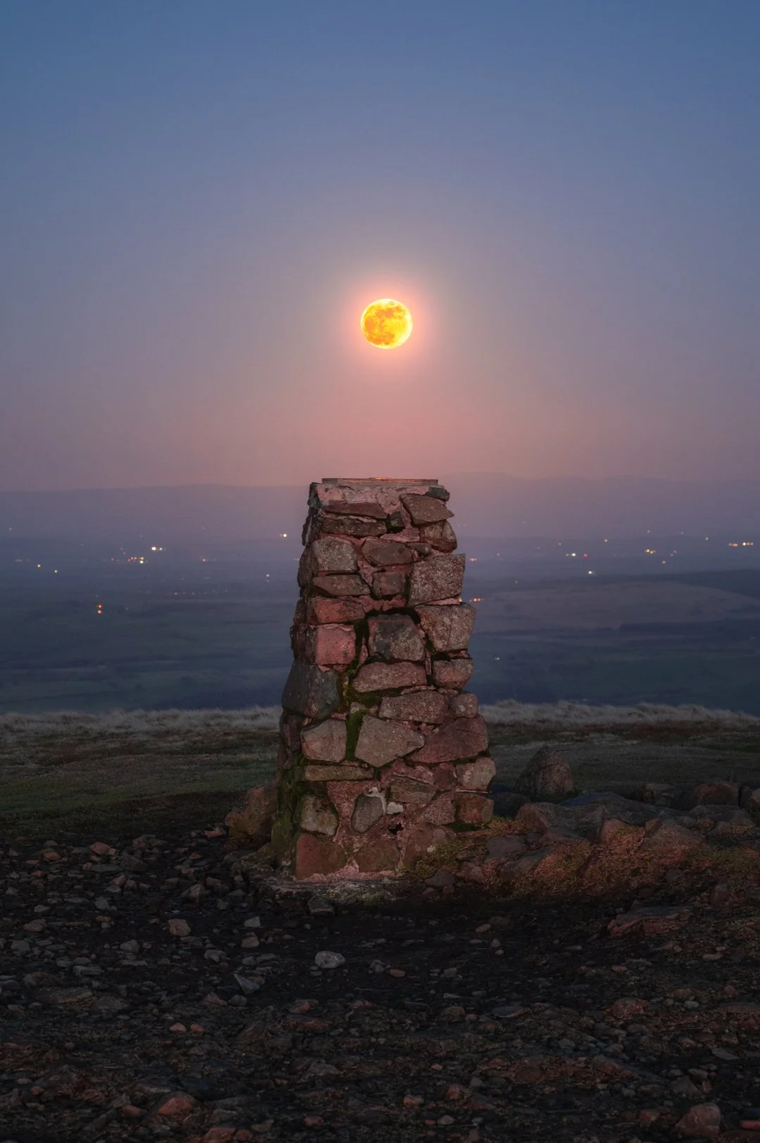 A stone cairn on a hilltop with a full moon above it, with a distant landscape and a clear evening sky, Lake District.