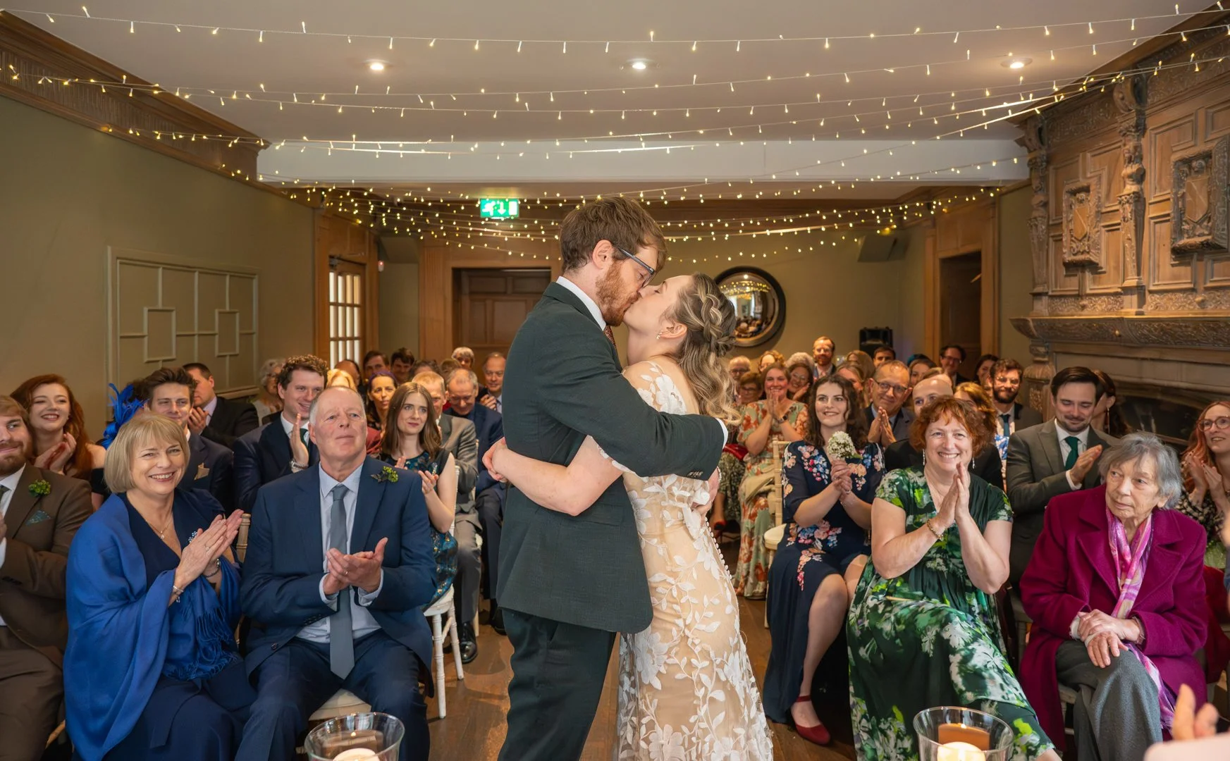 A bride and groom kiss while dancing during their wedding reception, with guests applauding and smiling around them in a decorated room with string lights and a fireplace, , peak district wedding photography. 