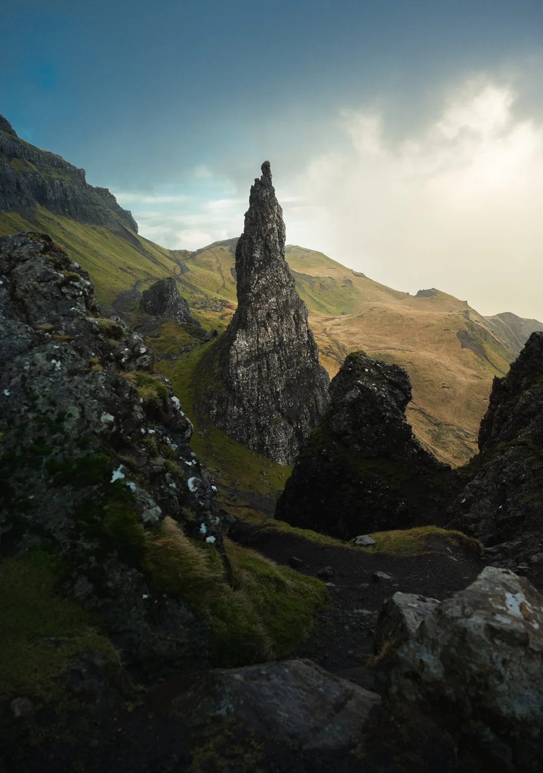 A rugged mountain landscape with a tall, narrow rock formation in the center surrounded by moss-covered rocks and grassy slopes, under a partly cloudy sky, Isle of Skye.