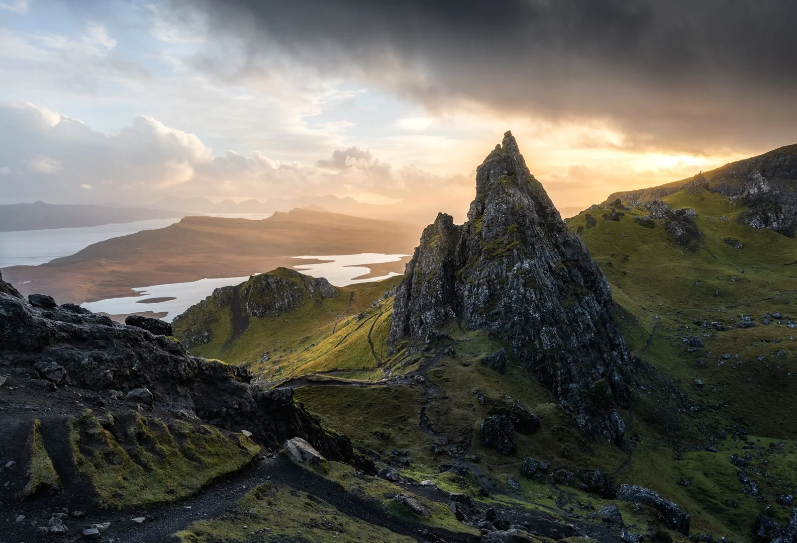 Scenic landscape of rocky mountain peaks with green slopes, a winding trail, lakes, and a dramatic cloudy sunset sky, Isle of Skye.
