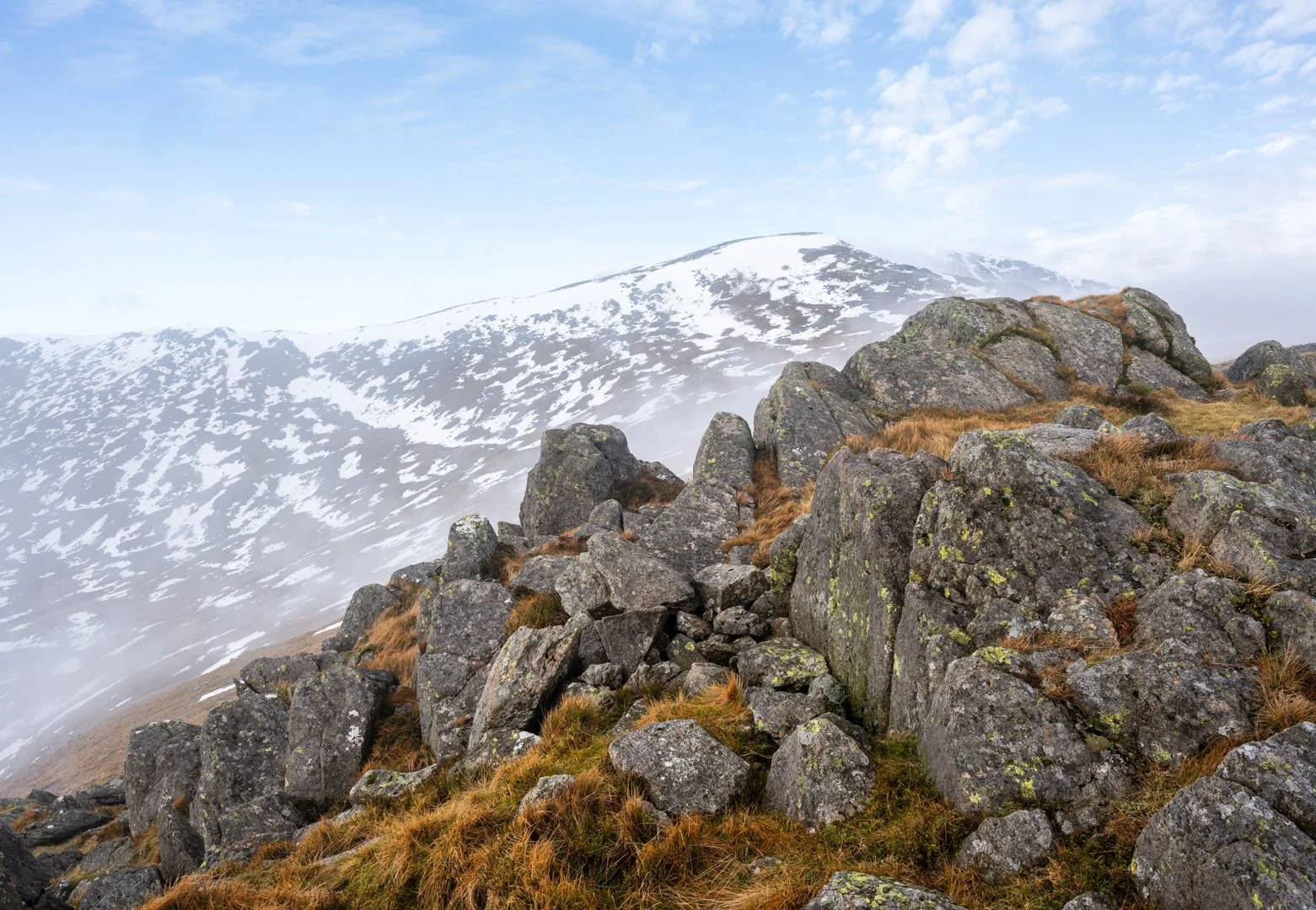 A rugged mountainous landscape with large rocks and patches of grass in the foreground, snow-covered peaks in the background, and a partly cloudy sky, Snowdonia. 