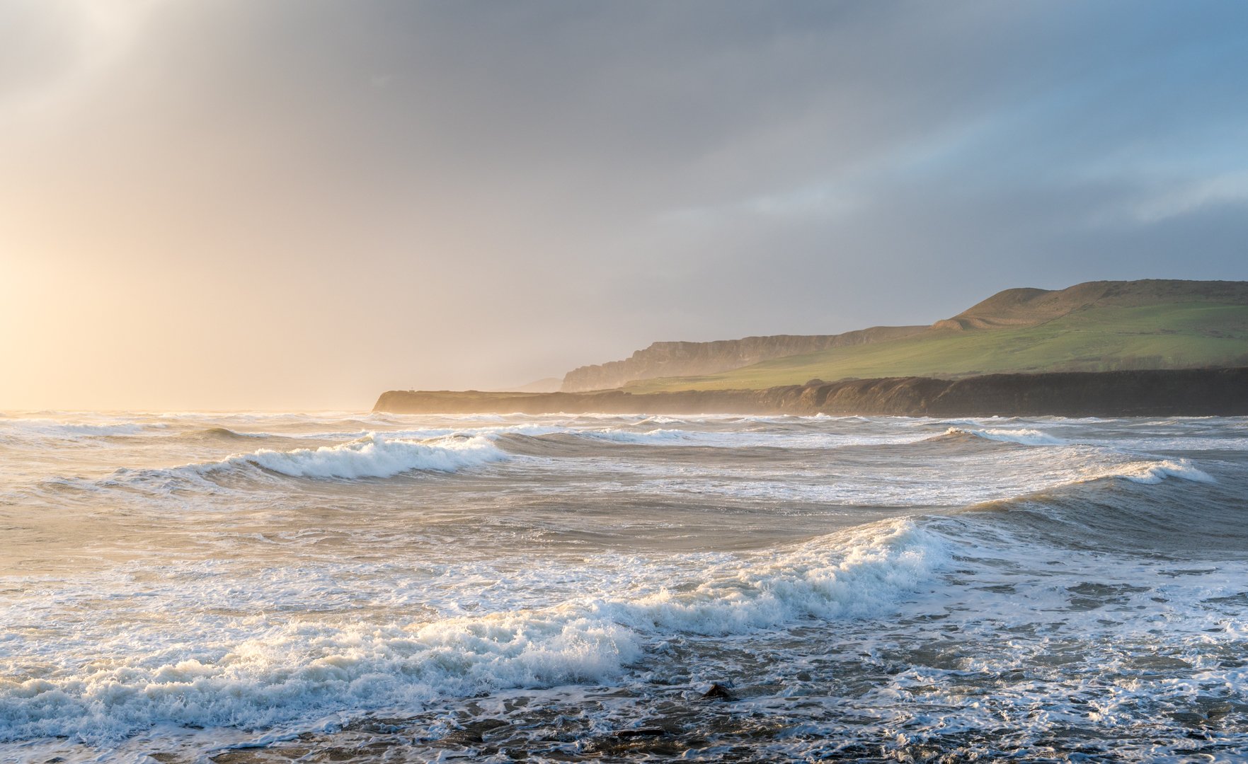Waves crashing on a rocky beach with green cliffs and hills in the background during sunset, Kimmeridge Bay