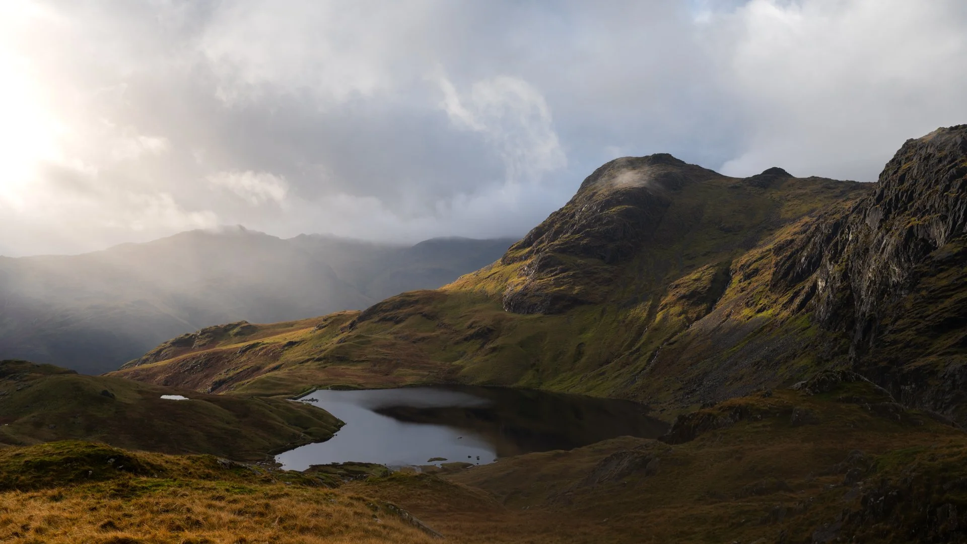 Mountain landscape with a small lake, cloudy sky, and sunlight breaking through clouds, Lake District.