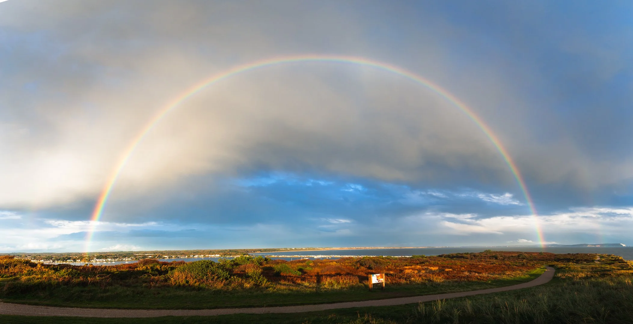 A vibrant rainbow arches across a partly cloudy sky over a coastal landscape with green vegetation, a winding path, and distant water, Hengistbury Head. 