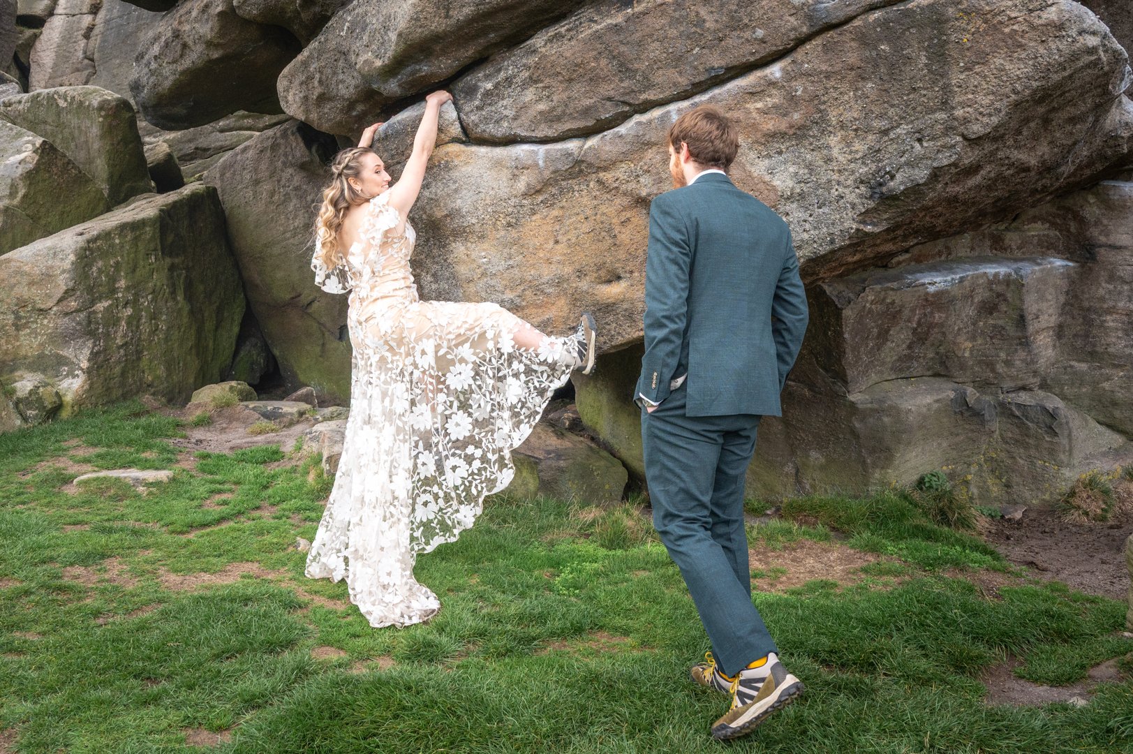 A woman in a long, floral lace dress climbing or leaning against a large rock formation, with a man in a suit watching her on a grassy outdoor area, peak district wedding photography. 