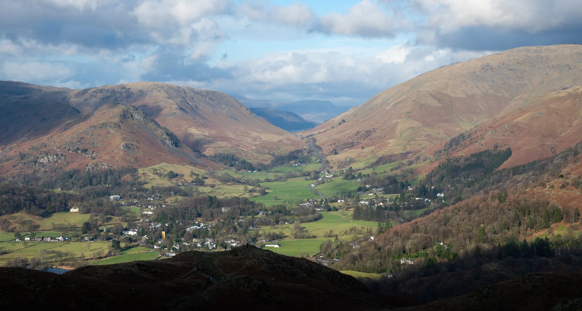 Scenic view of a valley with a small town surrounded by rolling green hills and mountains under a partly cloudy sky, Lake District.
