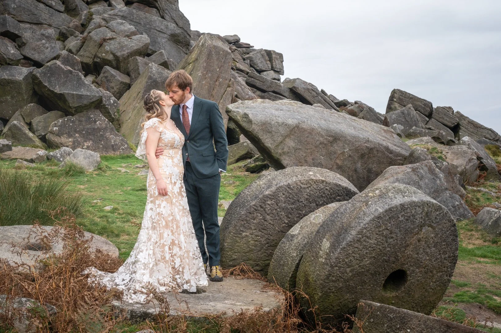 A couple kissing outdoors with large rocks in the background, woman in a floral gown and man in a suit, peak district wedding photography. 