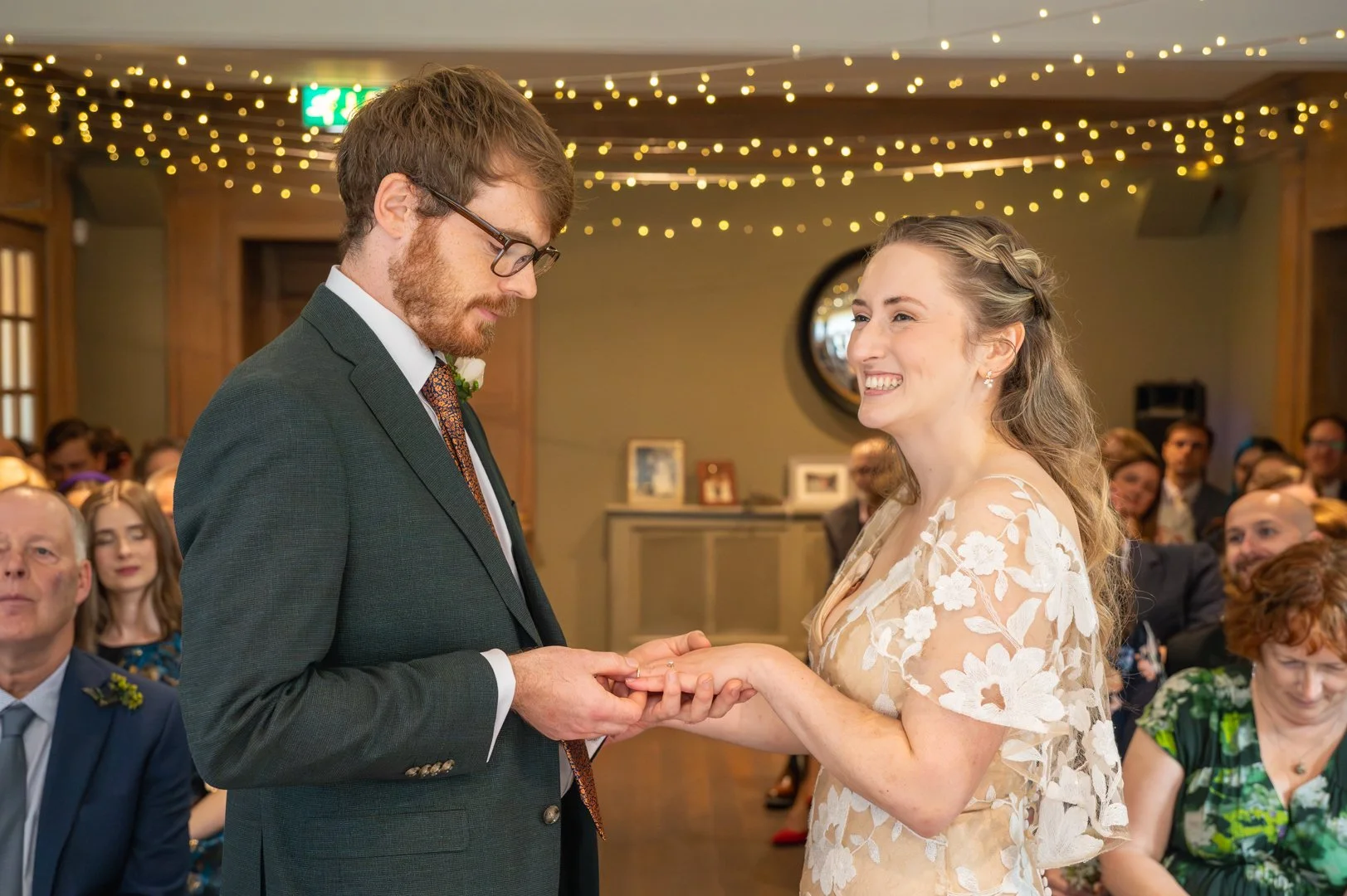 A couple exchanging rings during a wedding ceremony. The man is wearing a green suit and glasses, and the woman is smiling, wearing a white lace wedding dress with floral embroidery. Guests are seated in the background under string lights.