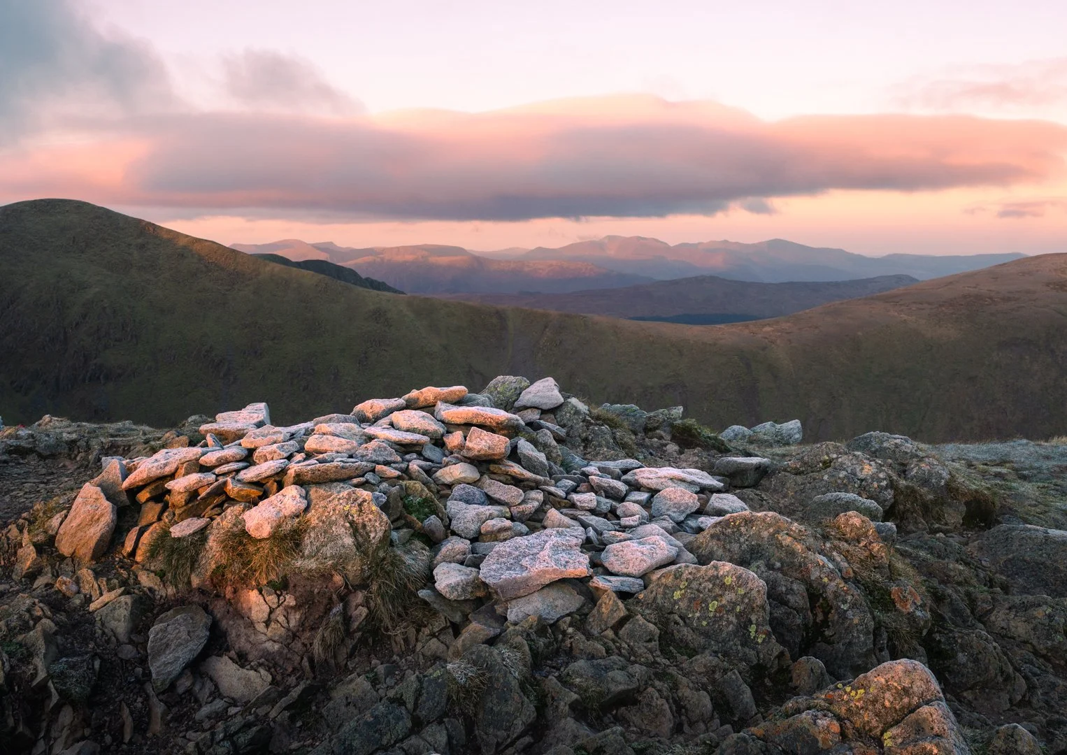 Scattered rocks and stones in a mountainous landscape during sunset or sunrise, with rolling green hills and a sky with pink and purple clouds, Lake District.