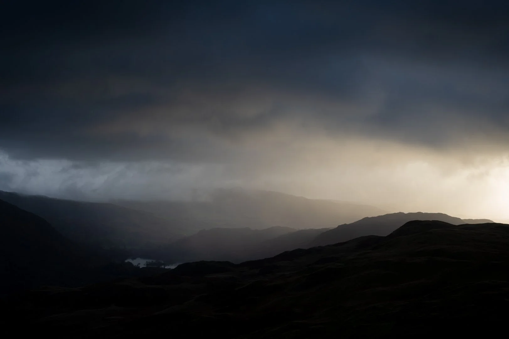 Dark storm clouds over a mountainous landscape with a faint light on the horizon, Lake District.