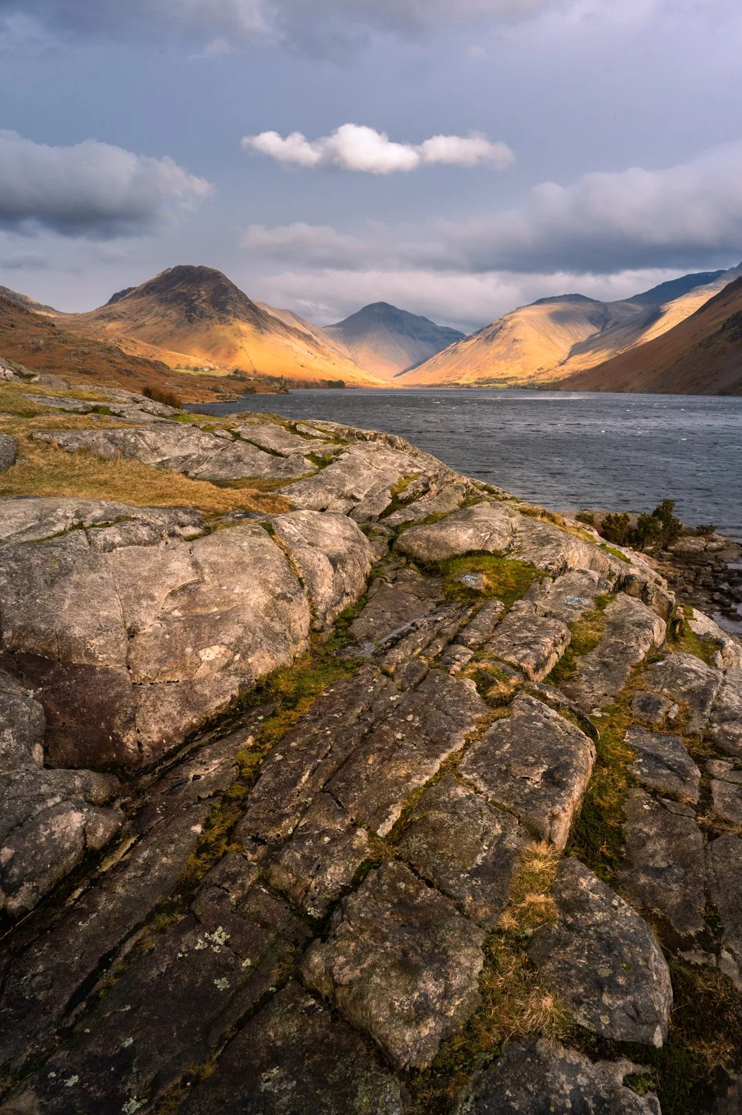 A scenic landscape of a lake surrounded by mountains and rocky terrain, with a partly cloudy sky overhead, Wast Water in the Lake District 