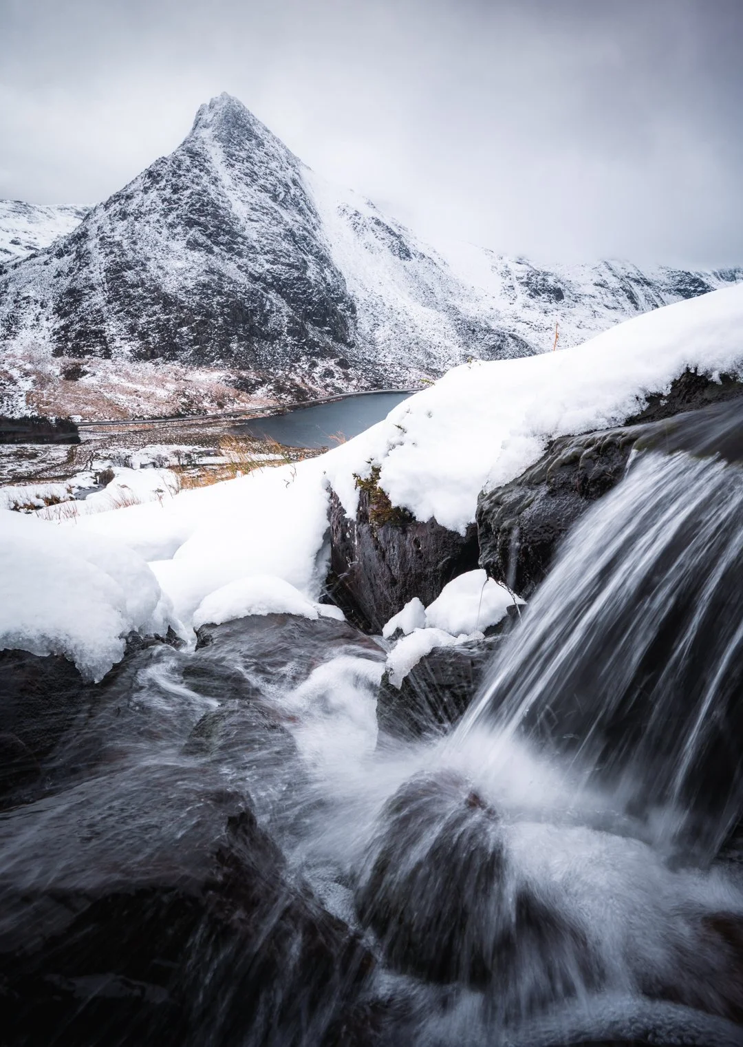 Snow-covered mountains with a river and waterfall in the foreground, overcast sky in the background, Tryfan Wales.