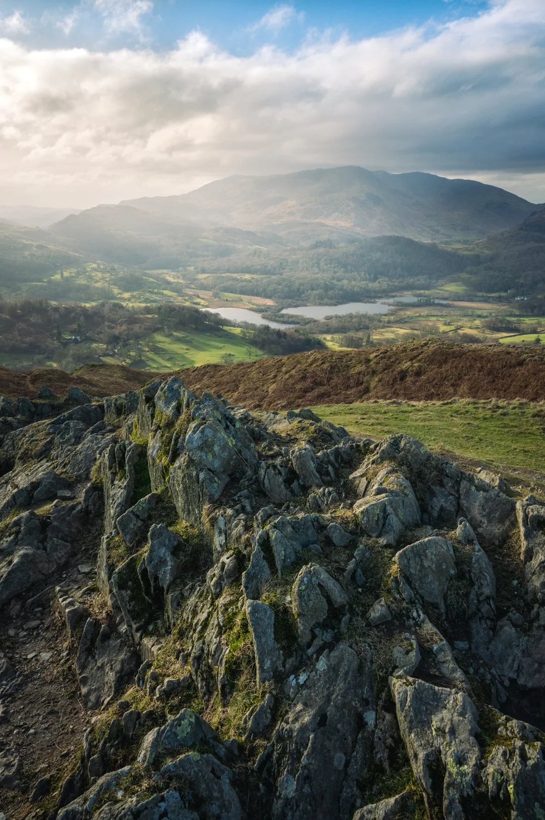 Rocky formation on a hillside overlooking a valley with a lake, green fields, and distant mountains under cloudy sky, Lake District.