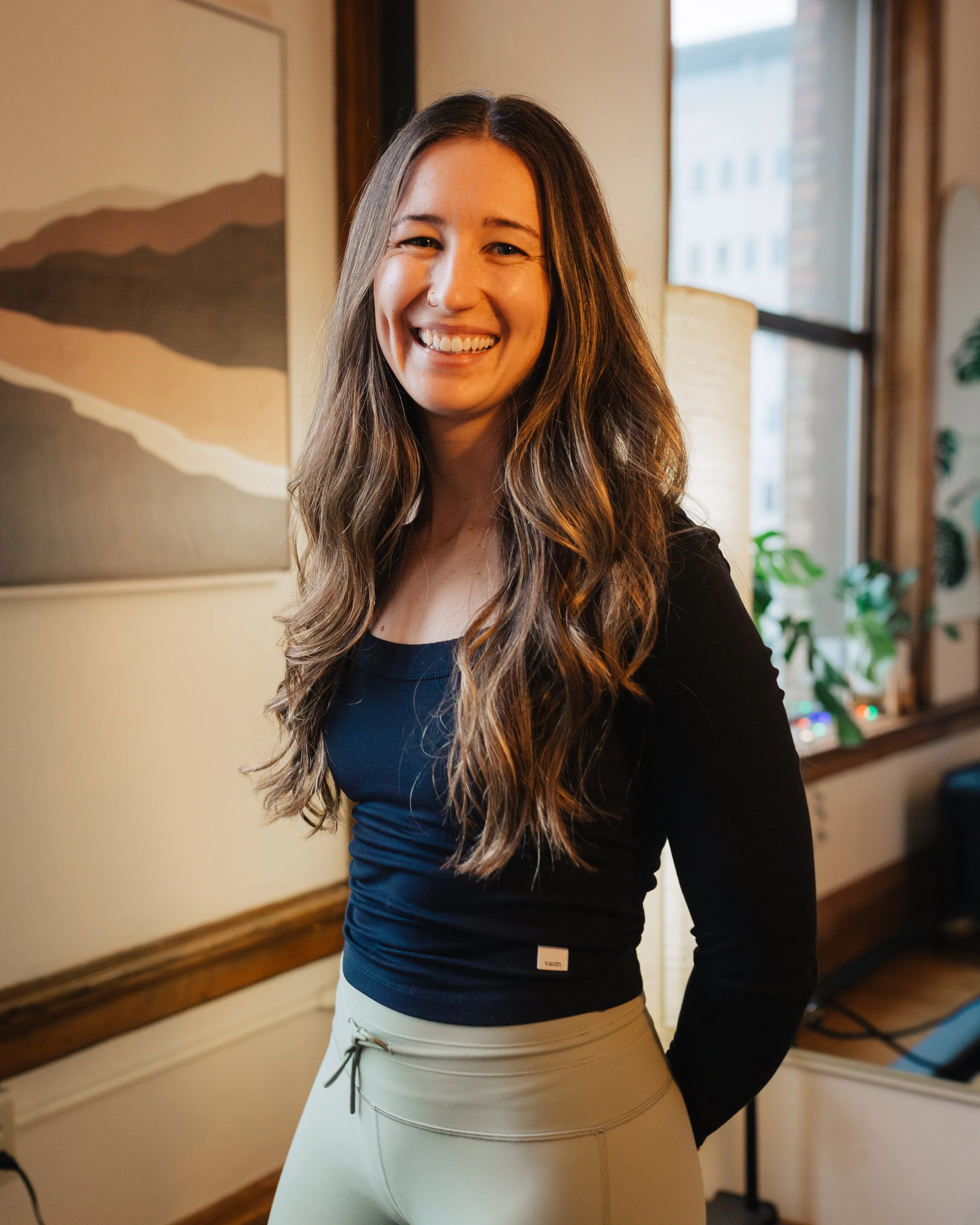 Olivia Crawford sitting on a black exercise mat on a wooden floor, smiling. She is wearing a beige sleeveless top, brown leggings, and white socks with black stripes. There is a red exercise ring near her foot, and gym equipment in the background.