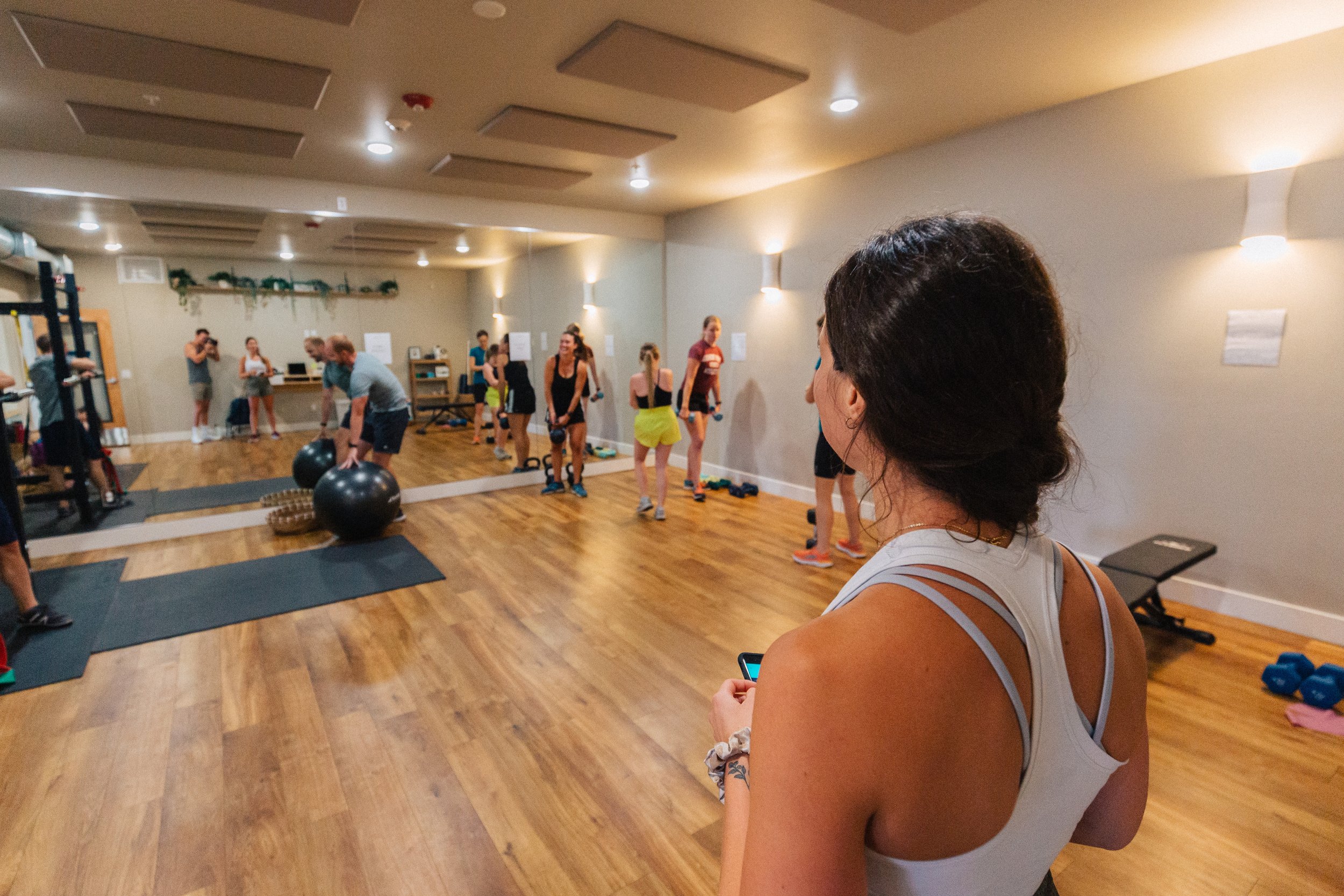 A group fitness class in a gym studio with participants lifting weights and using exercise balls, while a woman in the foreground checks her phone.