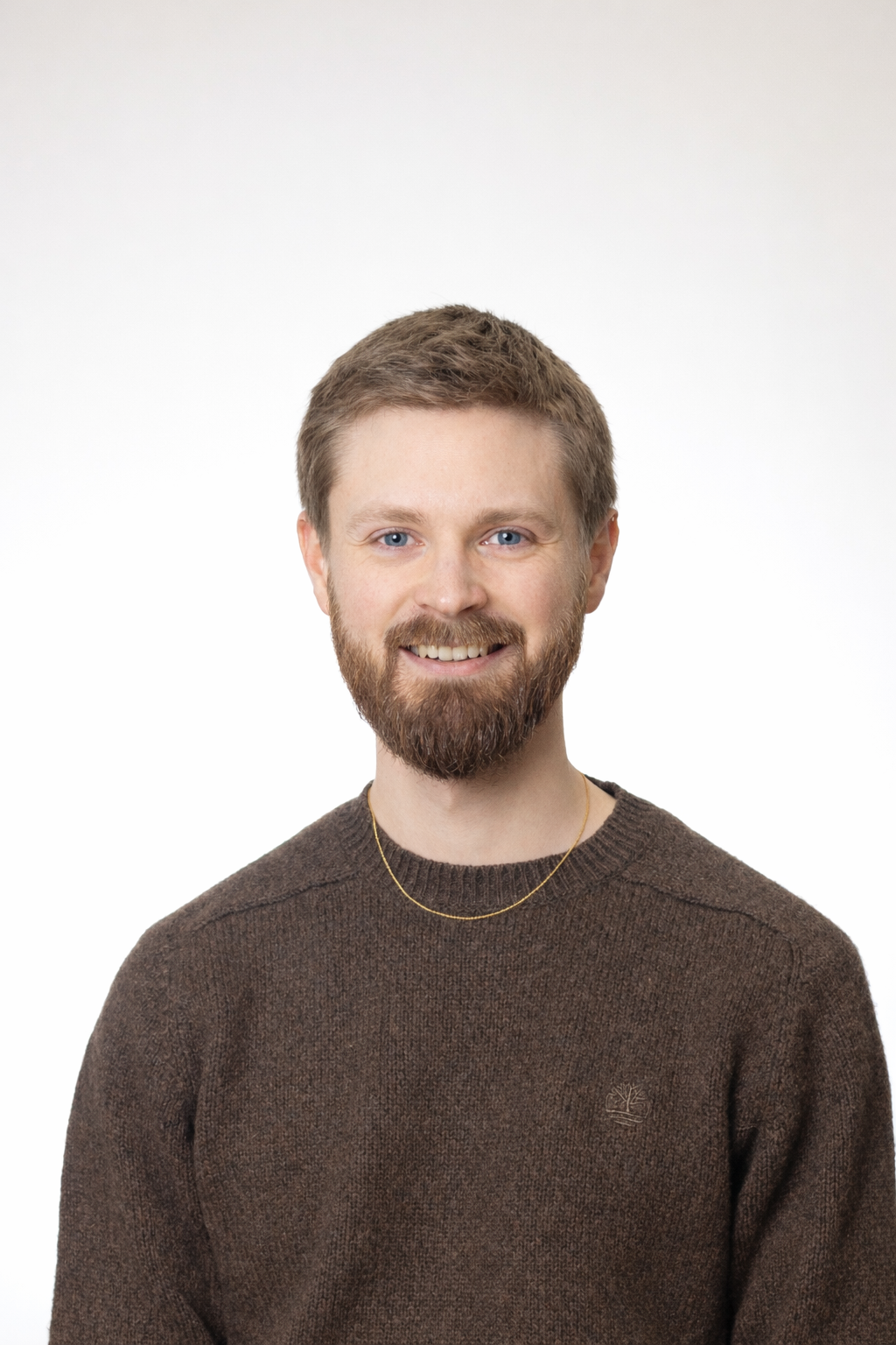 A young white man with short, light brown hair and a beard and glasses wears a grey humper