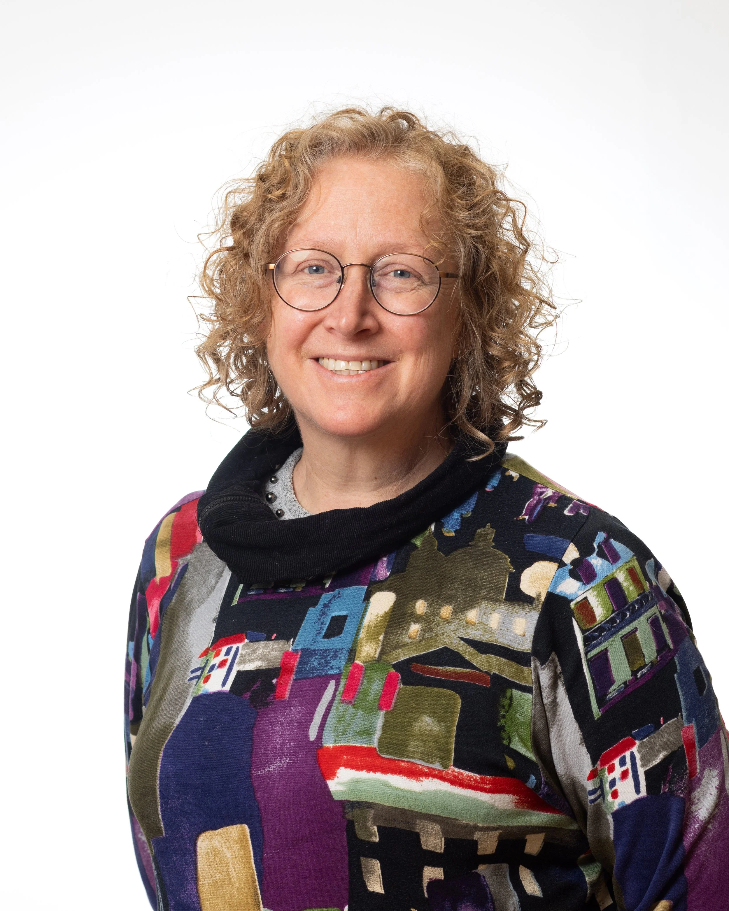 A white woman with curly chin-length blonde hair and black framed glasses smiles at the camera