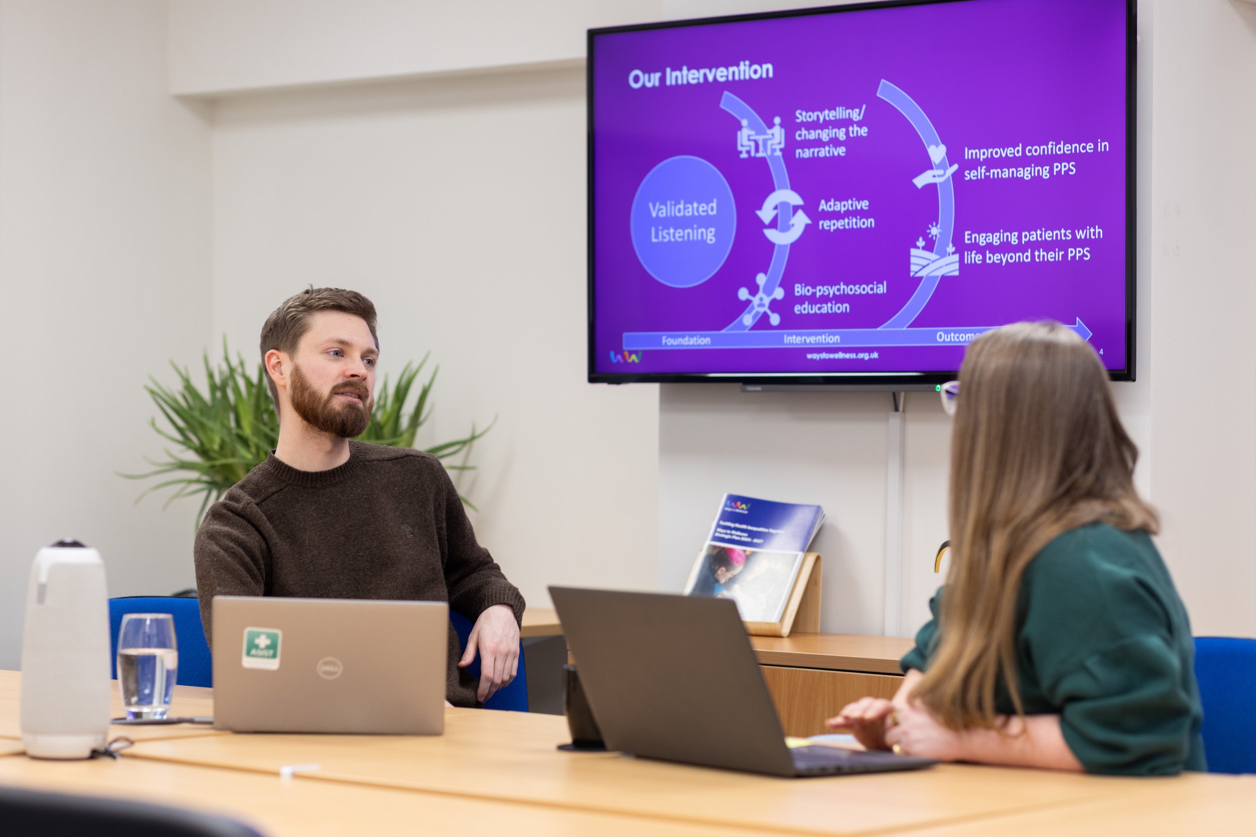 Two people sat at a desk with a presentation behind them.
