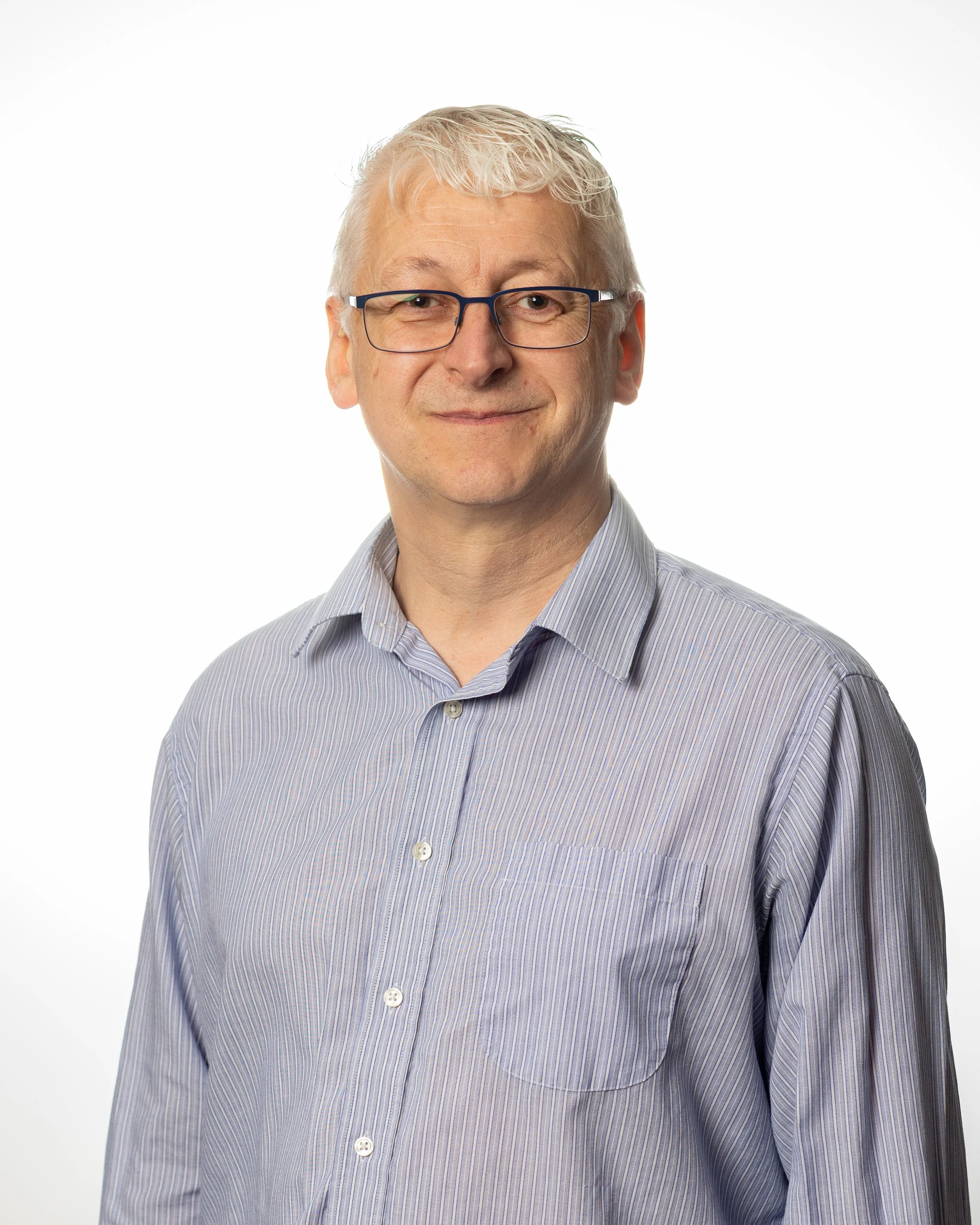 A white haired caucasian man with glasses smiles at the camera. He is wearing a pale blue shirt with an open collar