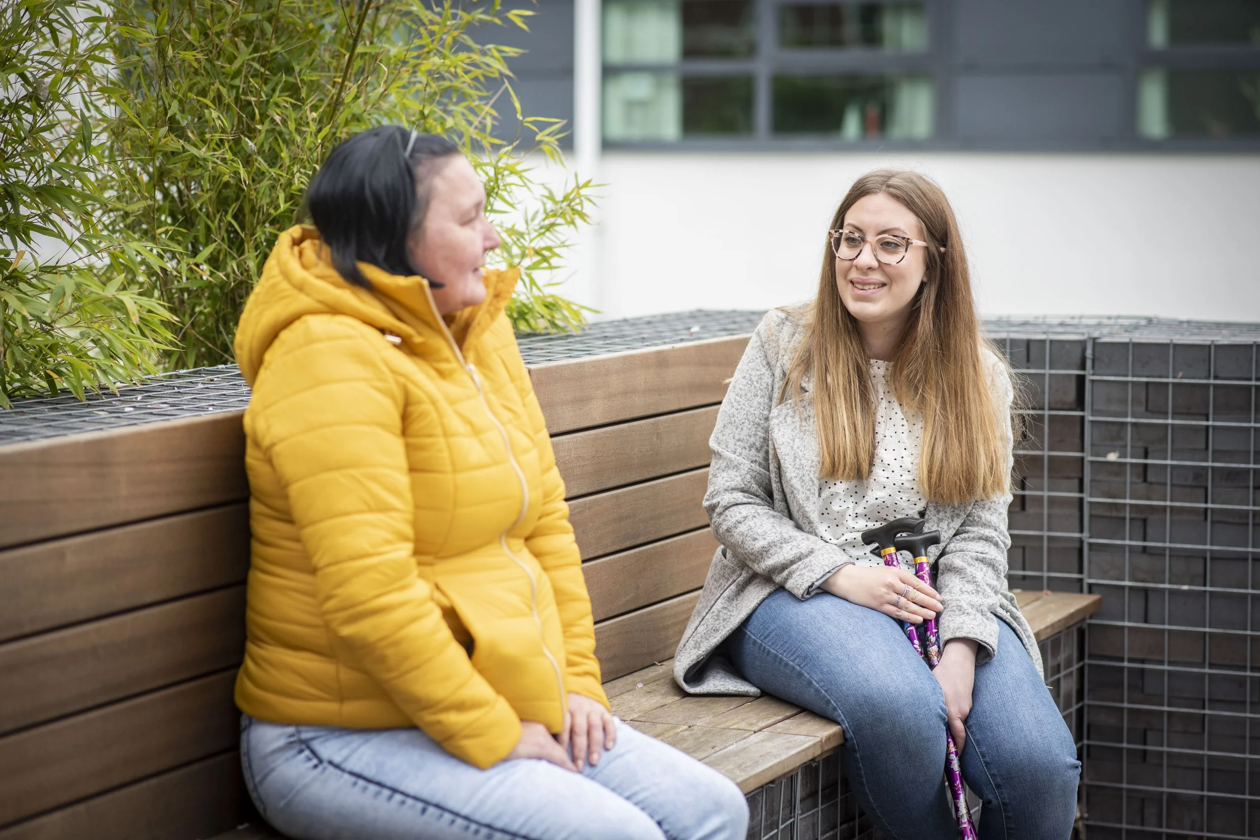 Two women sat on a bench talking. One is holding a walking stick.