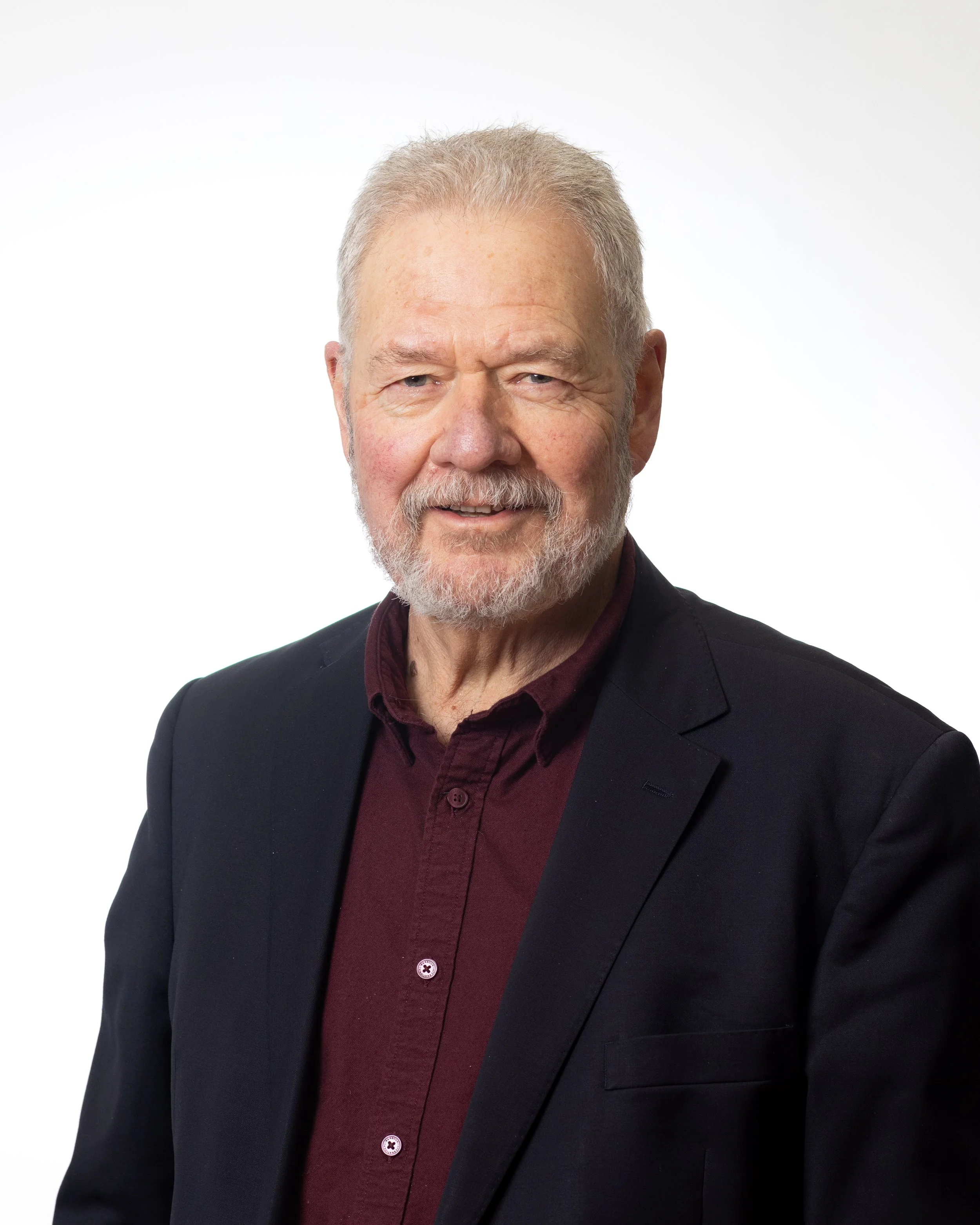 A middle aged man with short grey hair and beard, wearing a blue shirt and black jacket smiles to camera