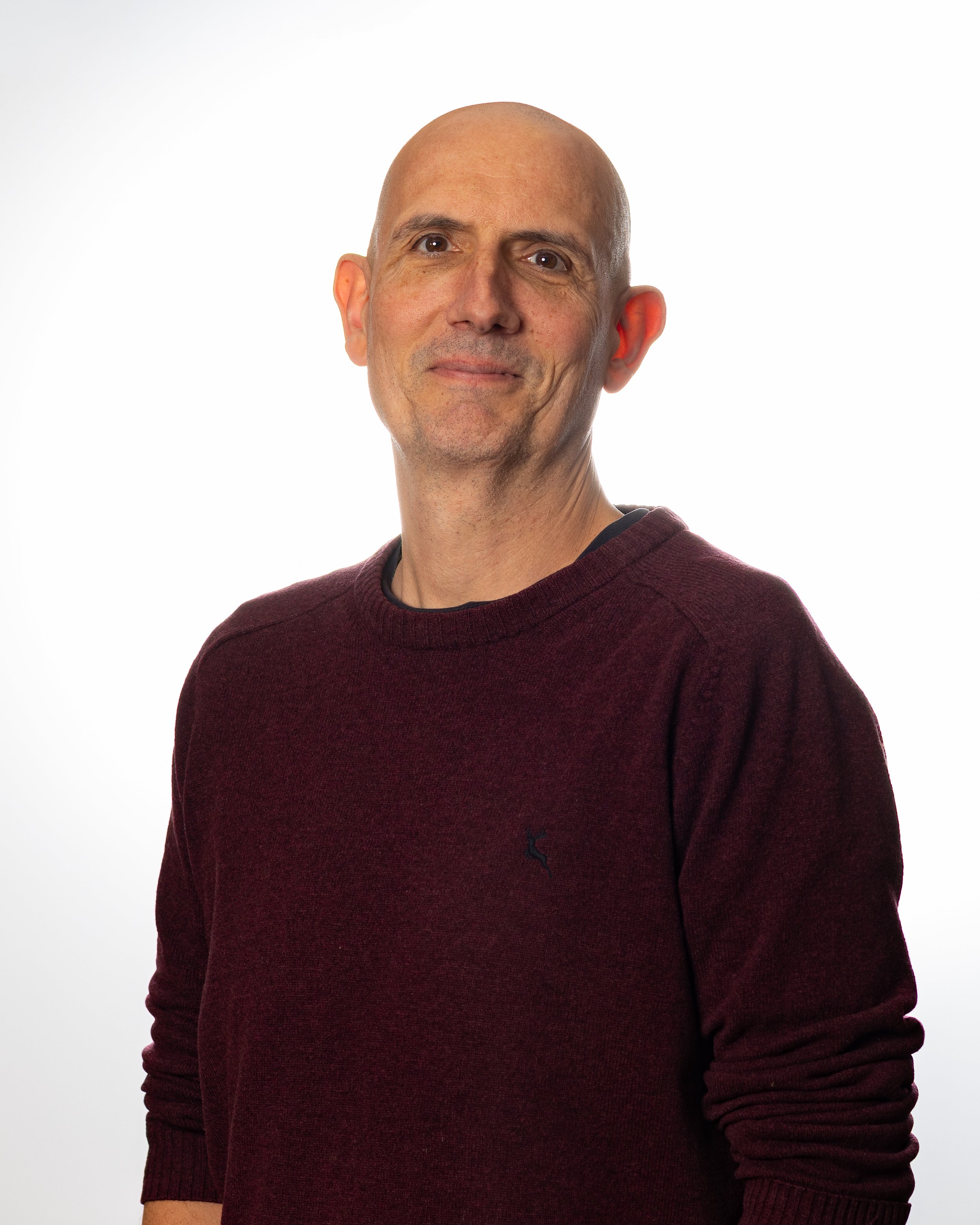 A tall, slim white man with no hair and a maroom coloured jumper smiles warmly without showing teeth