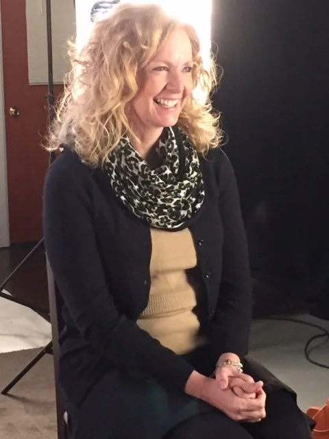 Woman smiling, wearing a black cardigan and patterned scarf, seated in a studio setting.